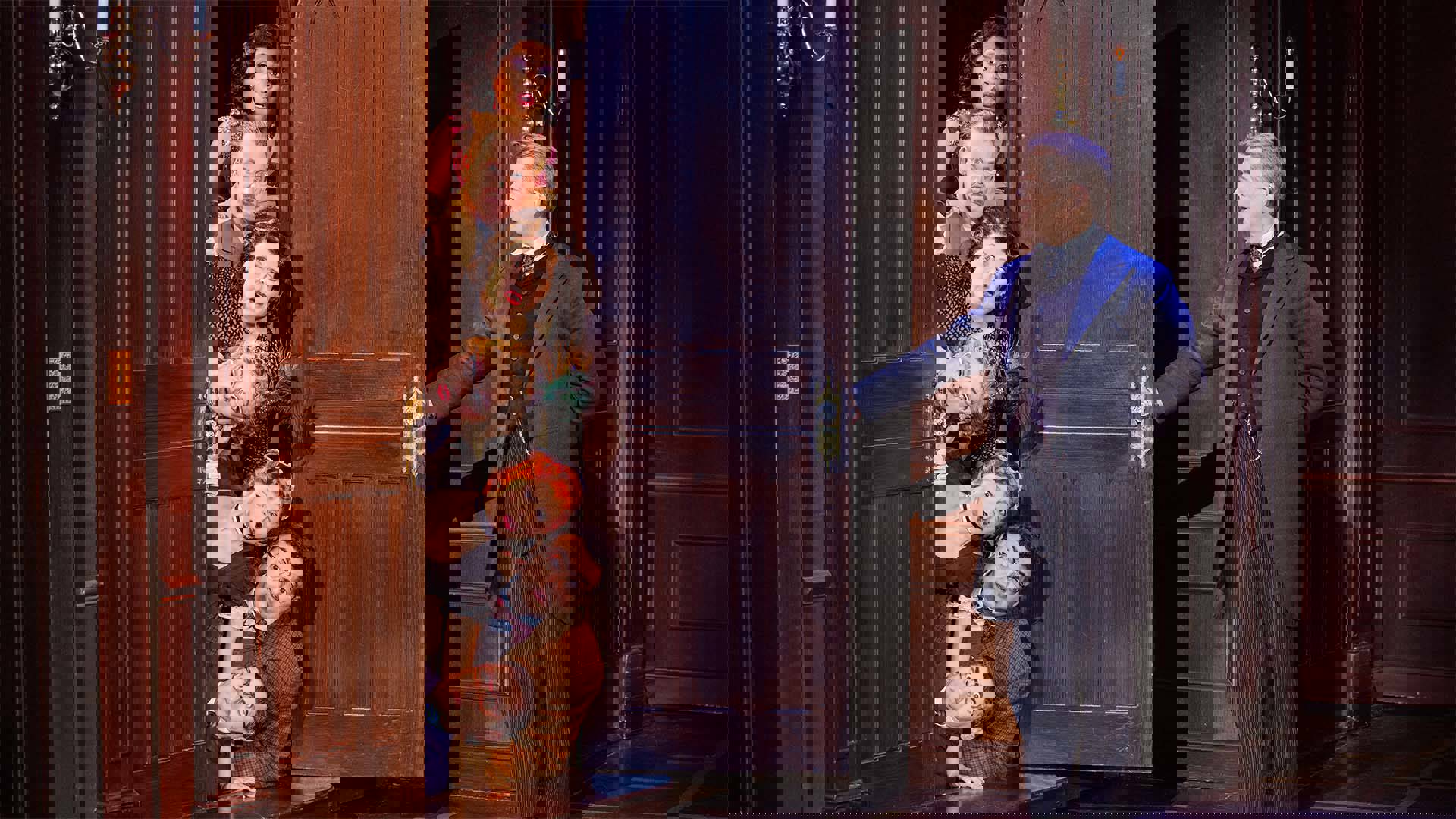 Seven actors lay stacked on top of each other horizontally, peeking out from behind an open door. Only their surprised faces are visible outside of the door frame. Across from them, a man in a suit has one arm outstretched, having just opened the door.