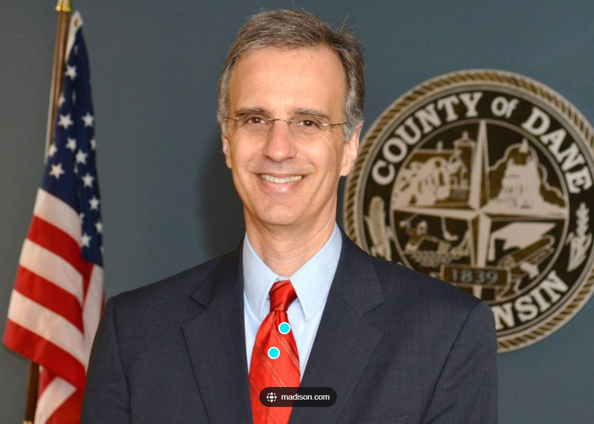 Headshot of a white man in a suit with dark eyes, short hair and glasses.