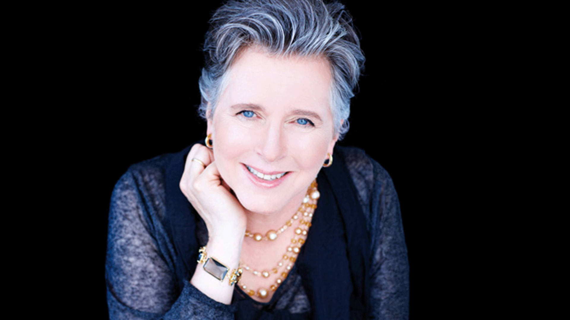 A black banner with a headshot of a middle aged white woman with short grey hair and blue eyes. She has a dark top and gold necklace on.