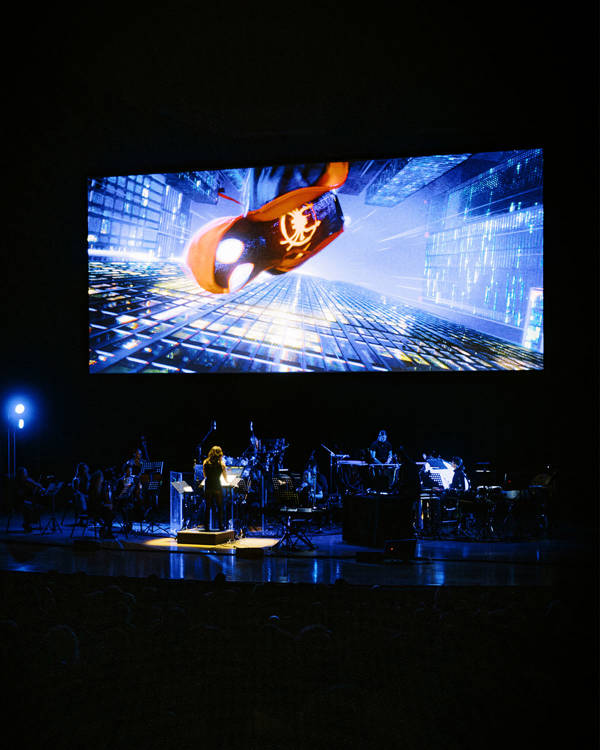 An orchestra on stage in dim blue light with a scene from spiderman into the spiderverse playing on a large screen.