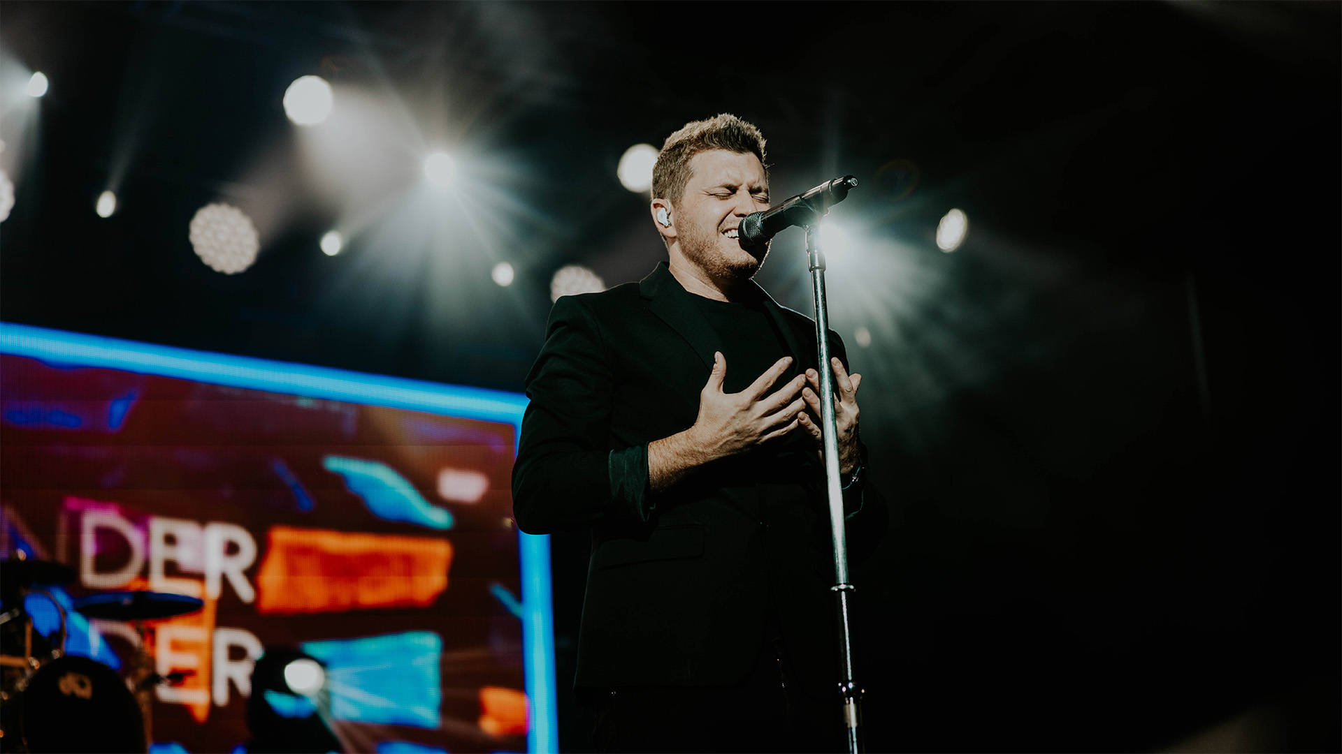 A man with light complexion and short hair in a dark outfit singing into a microphone.