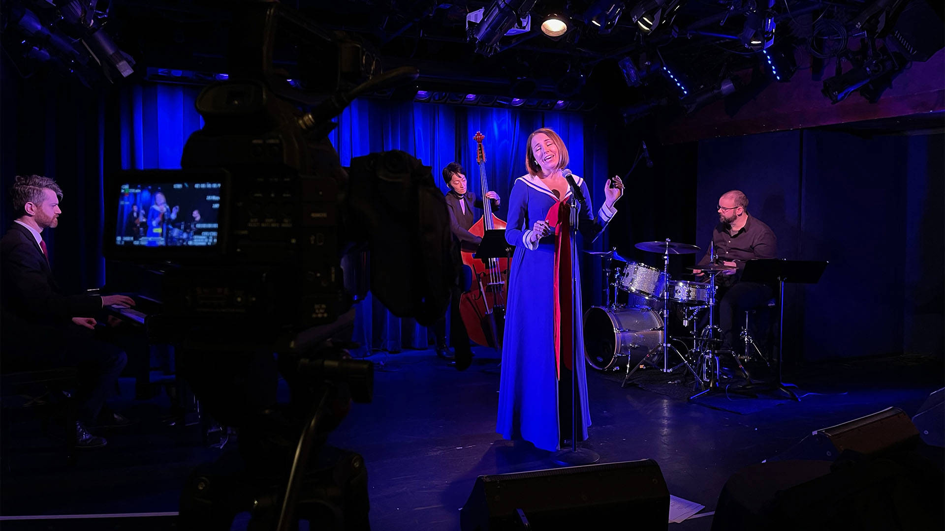 A caucasian woman singing in front of a band. In the foreground there is a tv camera capturing the performance. the woman has as blue outfit on, styled like a sailors outfit.