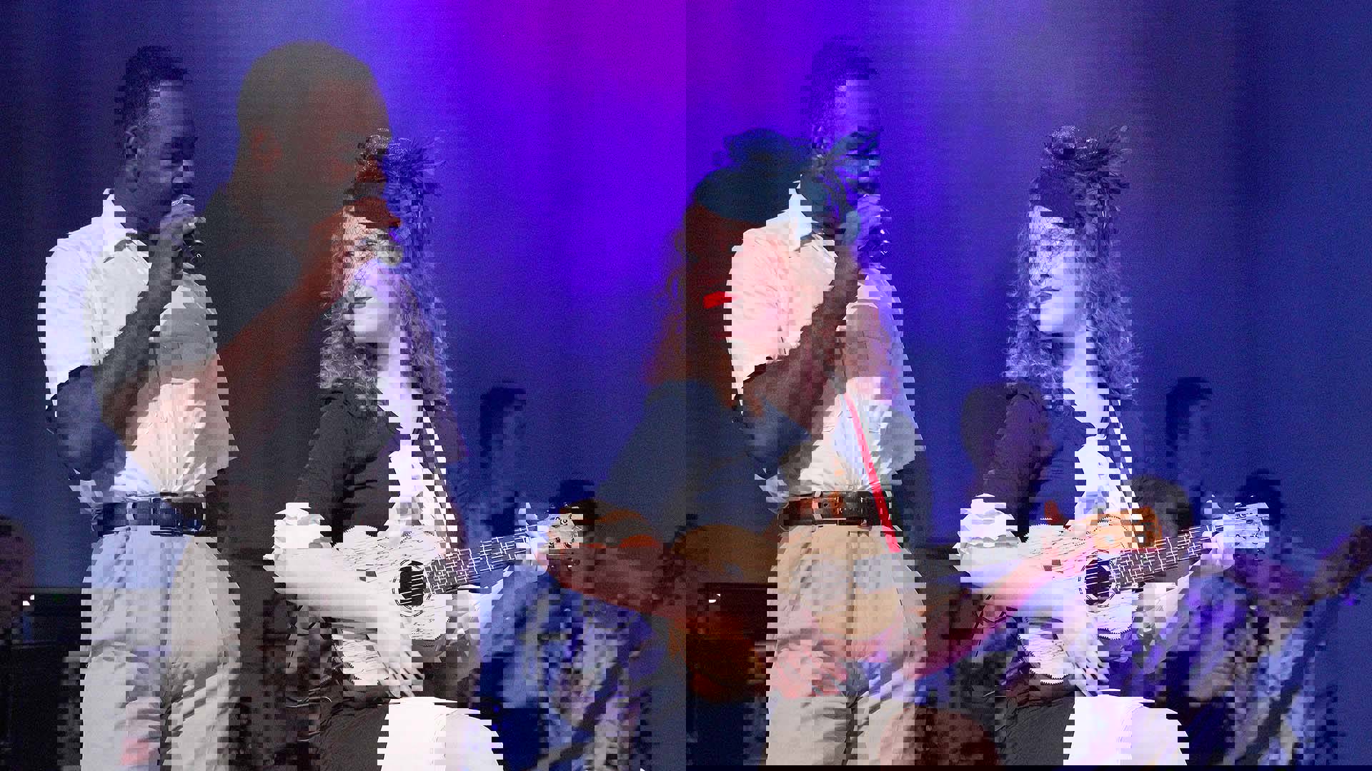 A young performer with dark complexion, short dark hair, a white shirt and khakis sings into a microphone next to another performer with light complexion and brown hair sitting on a stool holding a ukelel.