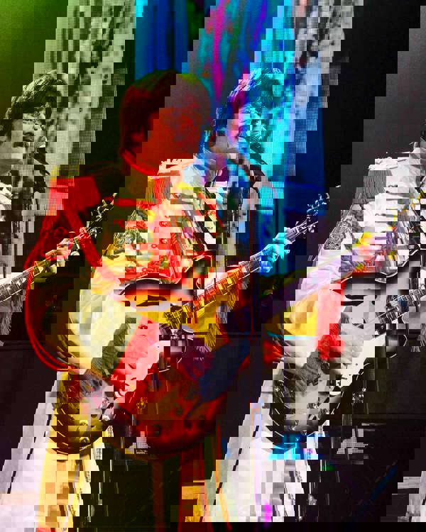 A John Lennon impersonator wearing a yellow Seargent Peppers outfit and holding a guitar. He has brown hair and a moustache.