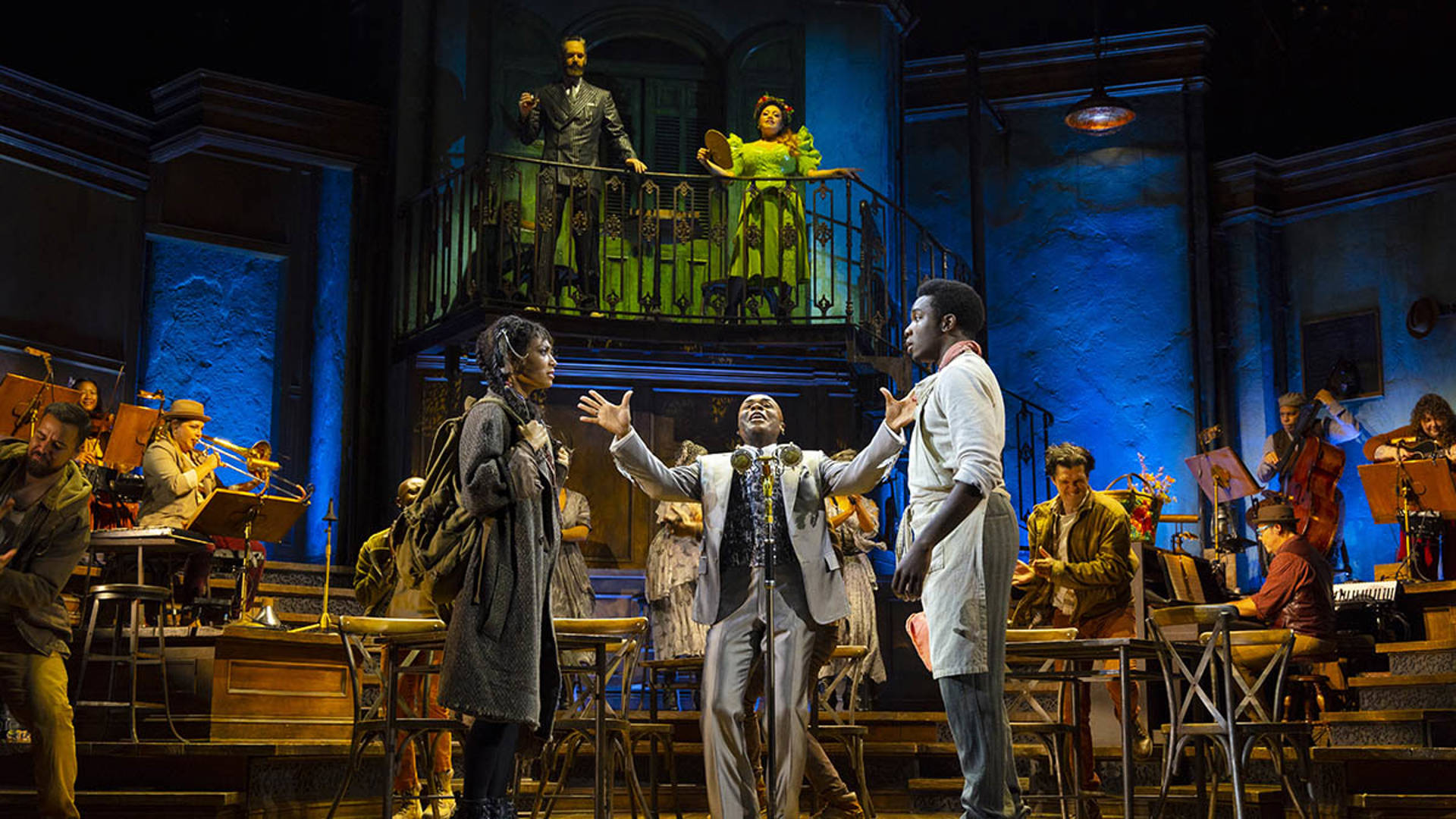 The cast of Hadestown performing on stage. At the center, an African American man and woman stare at eachother with straight faces, while another between them sings. He is wearing a silver suit and holding his hands up. 