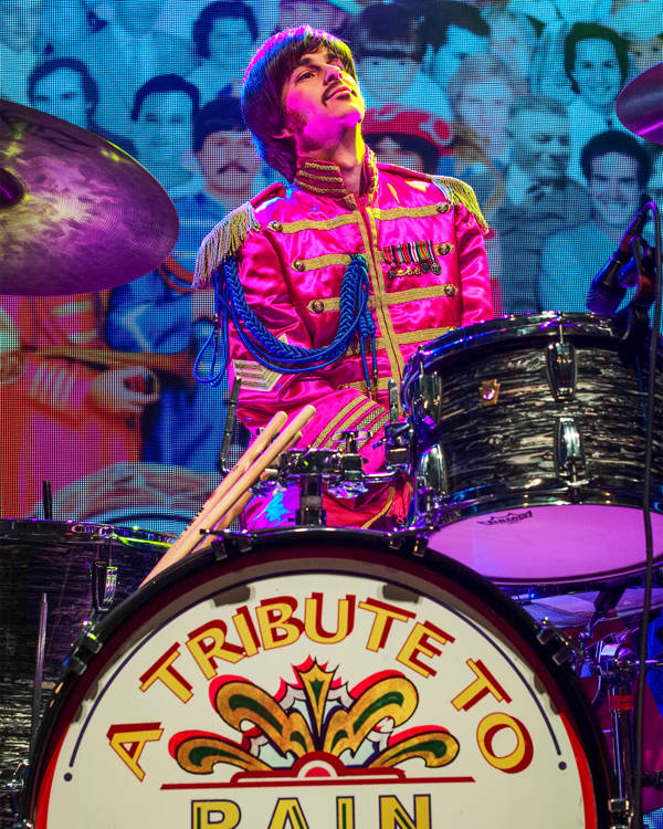 A Ringo Starr impersonator wearing a pink Seargent Peppers outfit and sitting at a drum set. He has brown hair and a moustache.