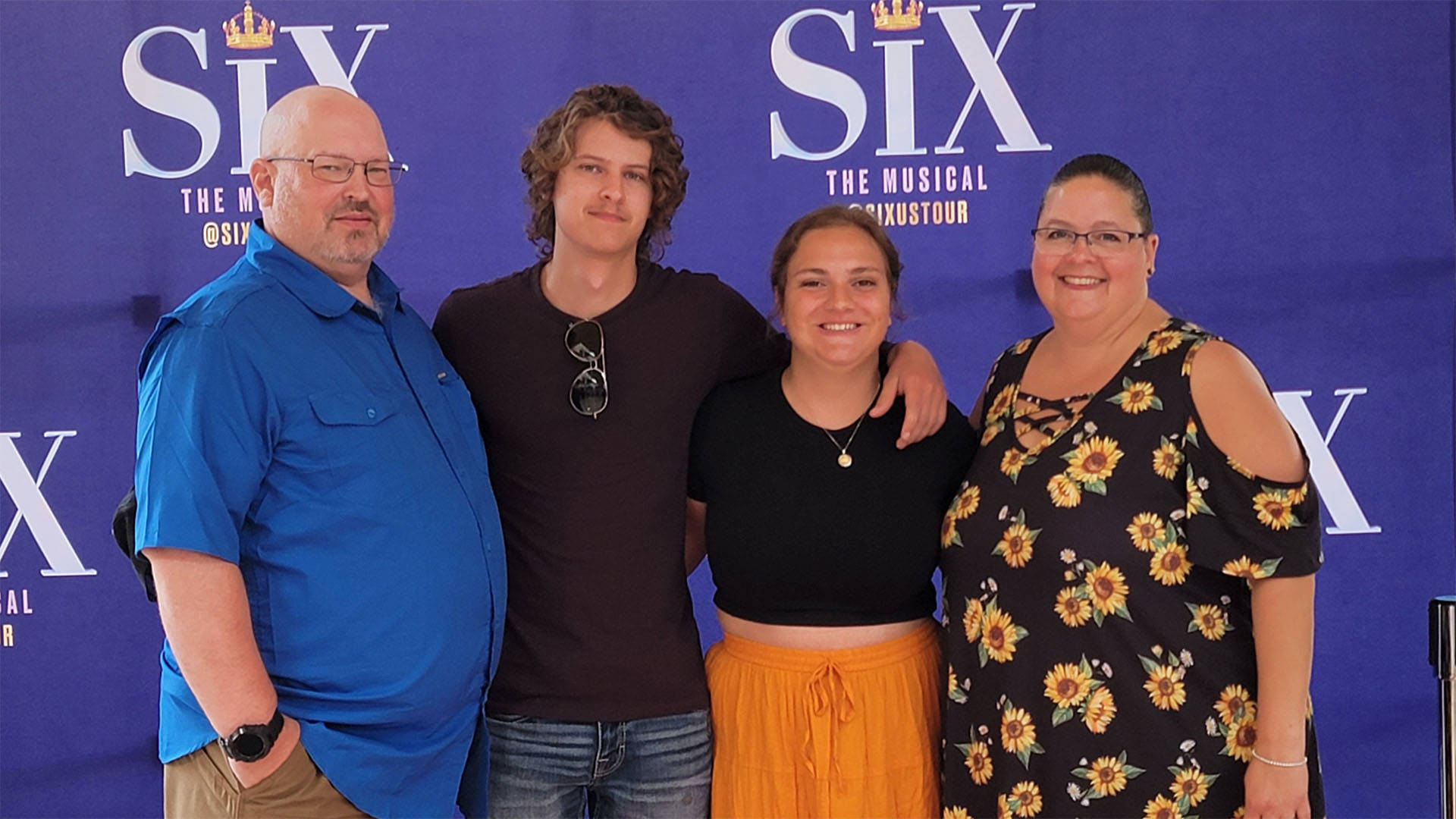Four people stand together for a photo in front of a purple backdrop the logo for the musical "SIX" repeating. The two on the outside are middle aged and the two on the inside are younger.
