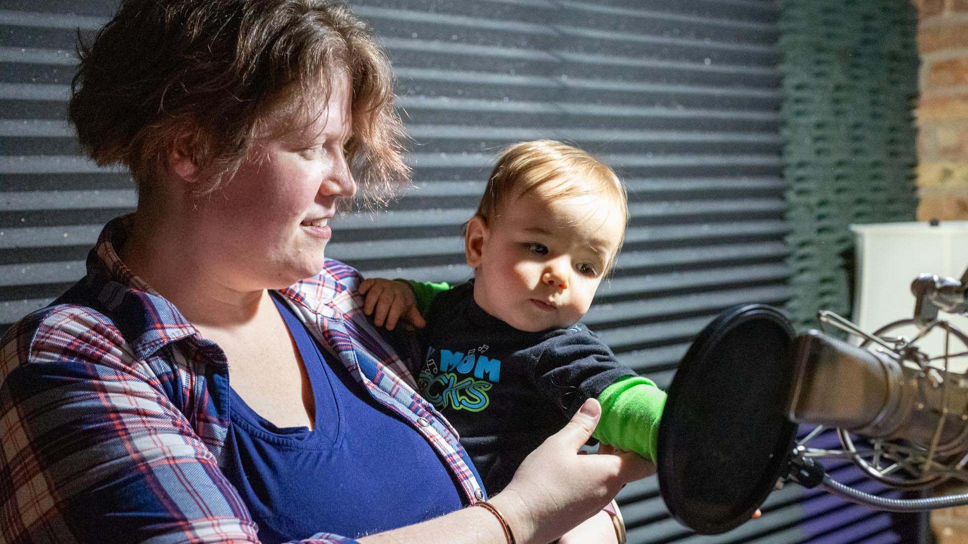 A woman with light complexion and medium length brown hair in a plaid shirt holding a baby touching a microphone