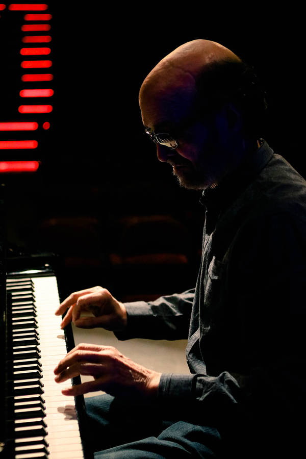 George Winston playing a piano. He is a caucasian man with a bald head, grey beard, and glasses
