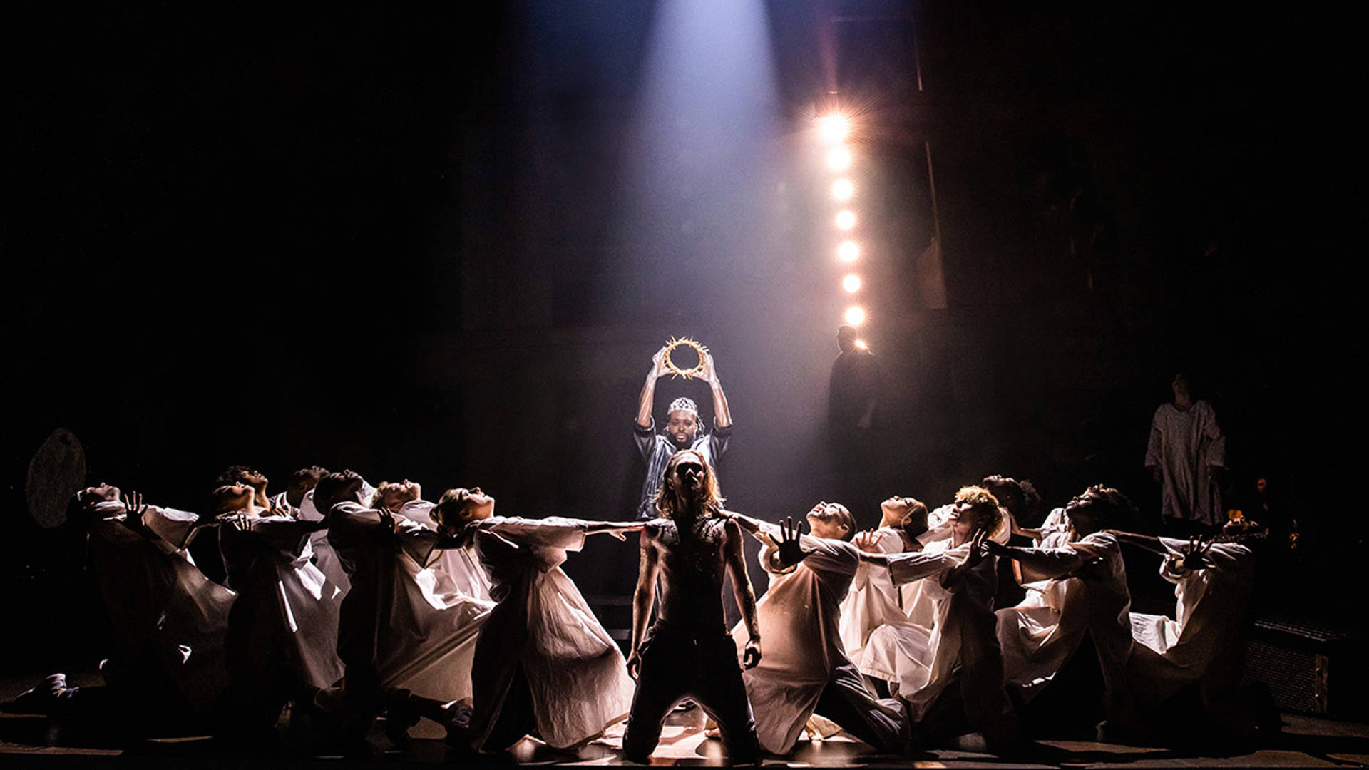 The cast of Jesus Christ Superstar kneel to the sides of a caucasian man as an African American man holds a crown of thorns behind him.