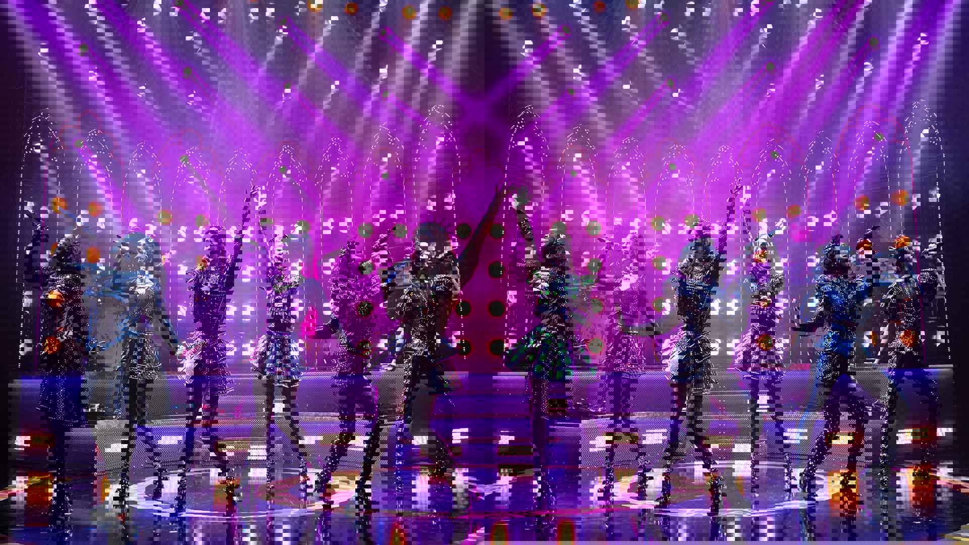 The cast of Six, in futuristic glittery outfits and holding microphones, performing on stage lit in purple.