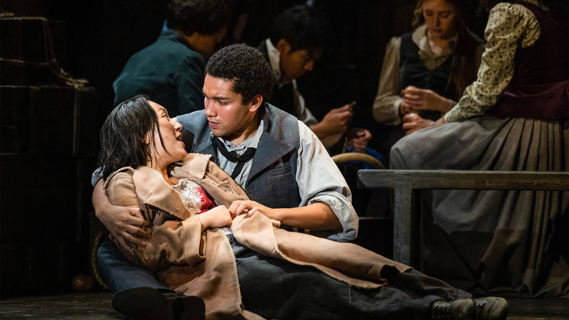 A man with dark short hair holds a woman in is arms on the ground. She has dark long hair and is wearing a long beige coat.