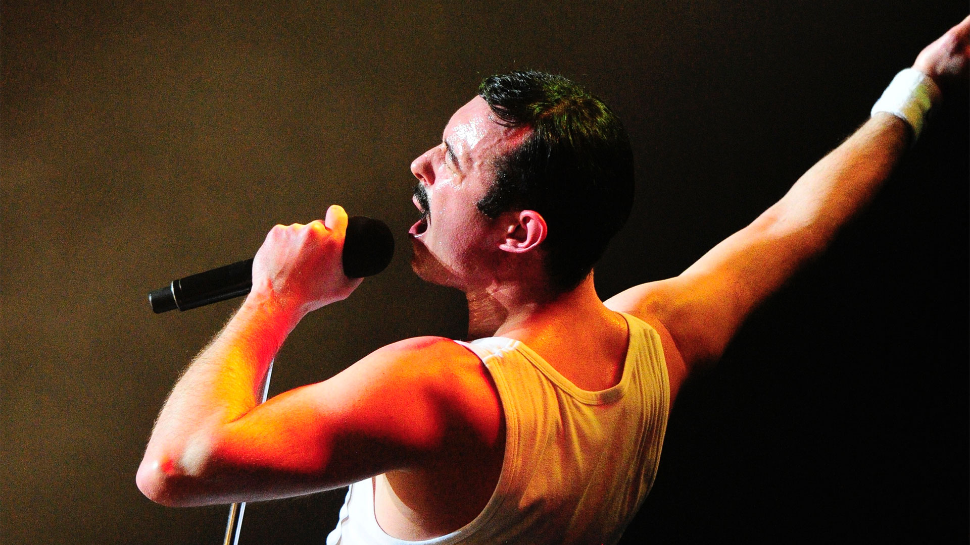 A caucasian man with short dark hair and moustache sings into a microphone with his right hand in the air. He is wearing a white tank top.
