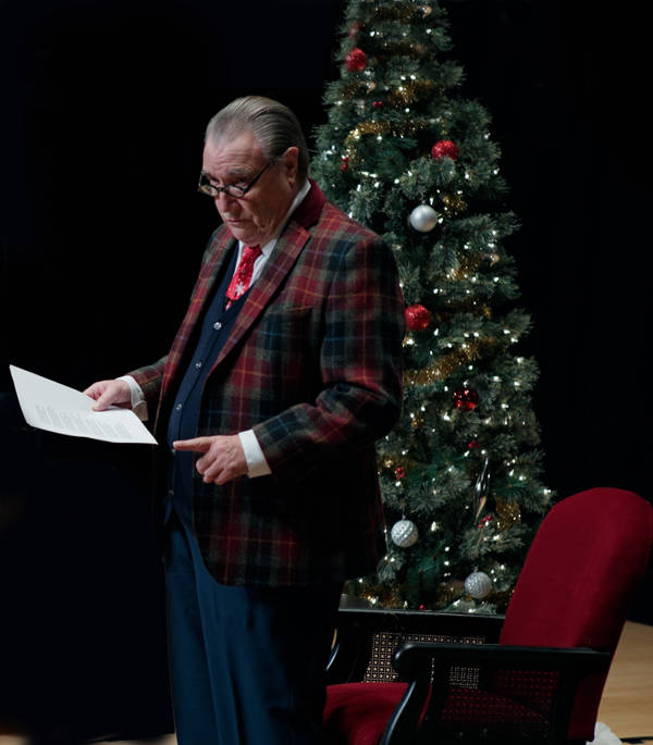 An older, white man with a receding hairline and black glasses in a blue suit and plaid jacket. He is standing and holding a sheet of paper in front of a Christmas tree.