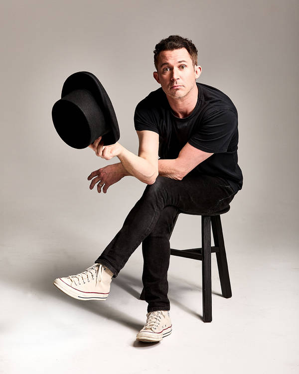 Justin Willman, a caucasian man with short brown hair and brown eyes, sitting ona black stool. He is wearing black jeans and a black tshirt. He is holing a black tophat in his right hand. His legs are crossed and he is looking towards the viewer.