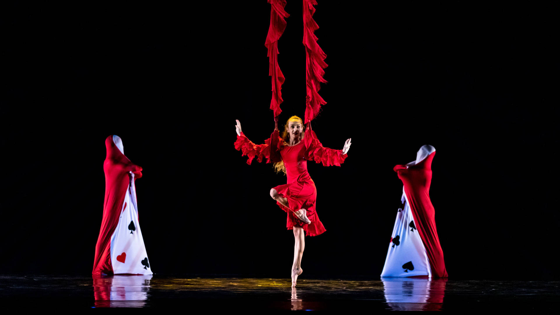 A dancer in a red flowing dress with long brown hair standing on one leg's tippy toes. There are two ruffly red fabric ropes at each shoulder slightly lifting her up. Two people in playing card costumes stand on either side.