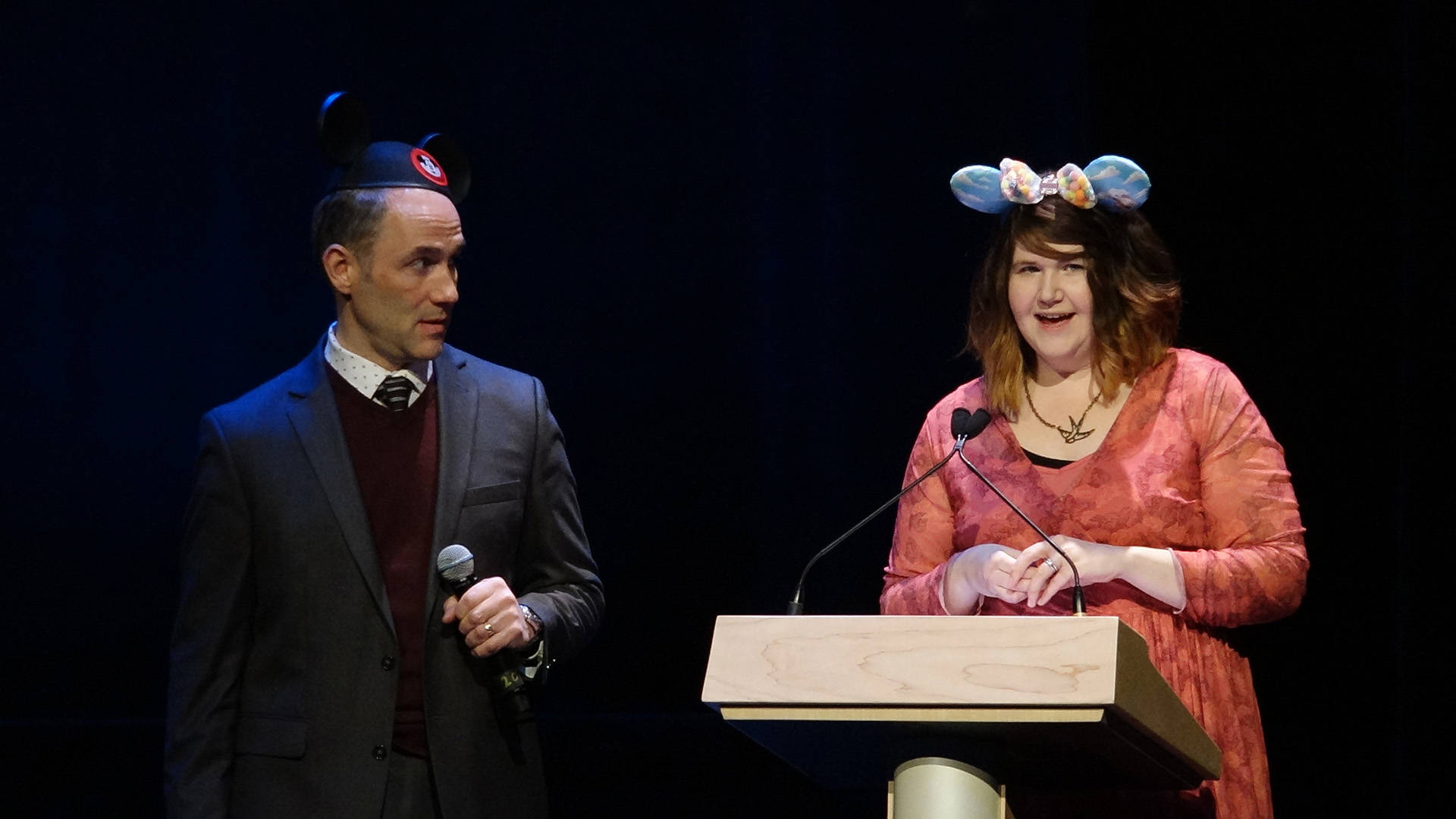A fair skinned man in a suit and Mickey Mouse hat holding a microphone standing next to a fair skinned woman behind of a podium in mouse ears.
