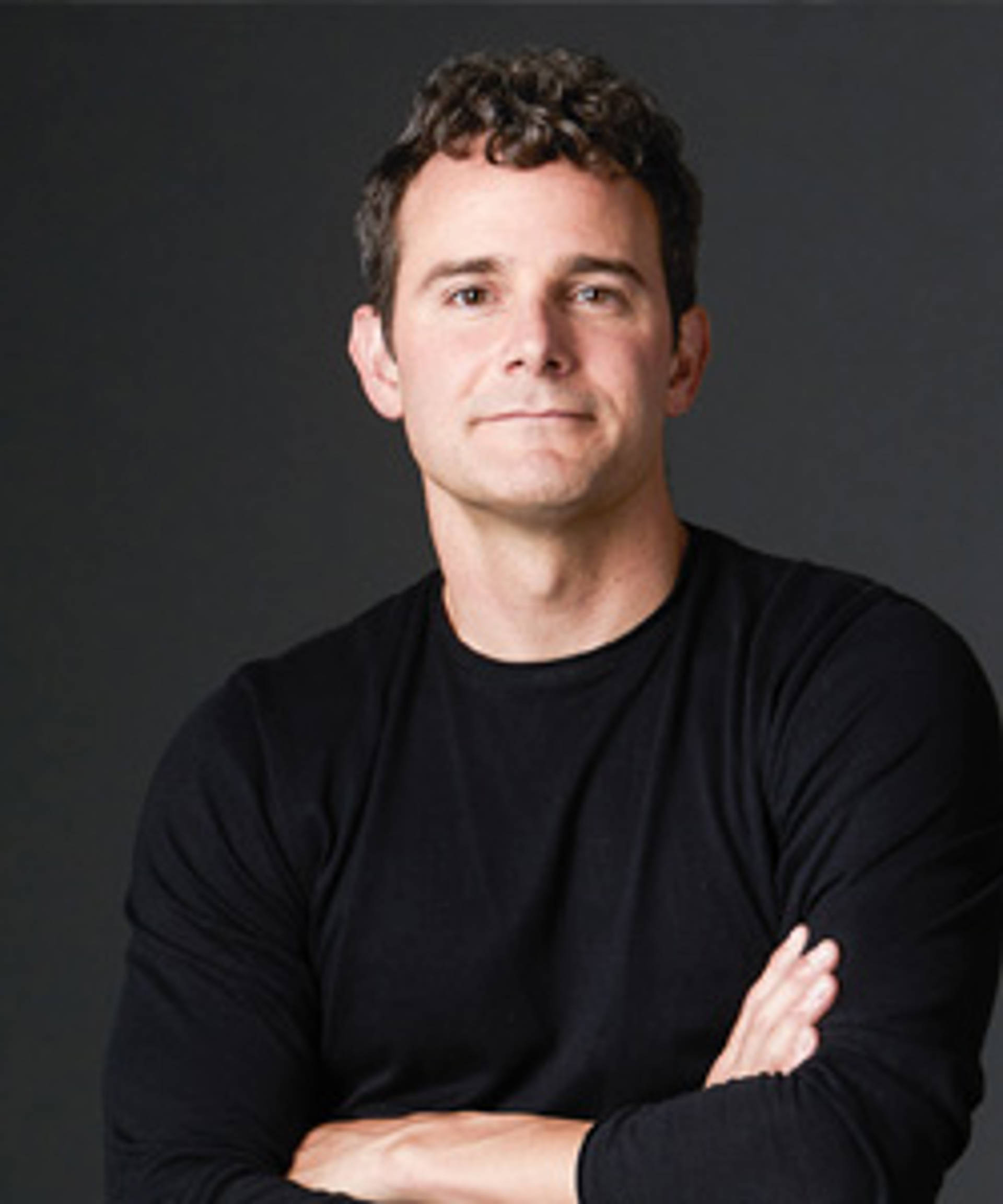 A man with light tone skin and a black top with short curly hair.