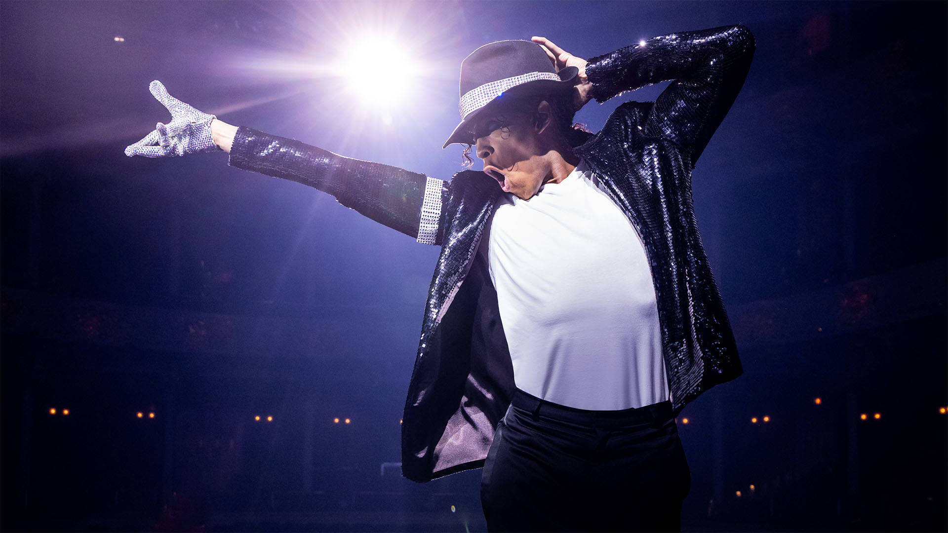 An actor playing Michael Jackson dancing on stage. He has medium complexion, dark hair, a dark jacket, hat, and a white shirt, and dark pants. There is a single spotlight on him.