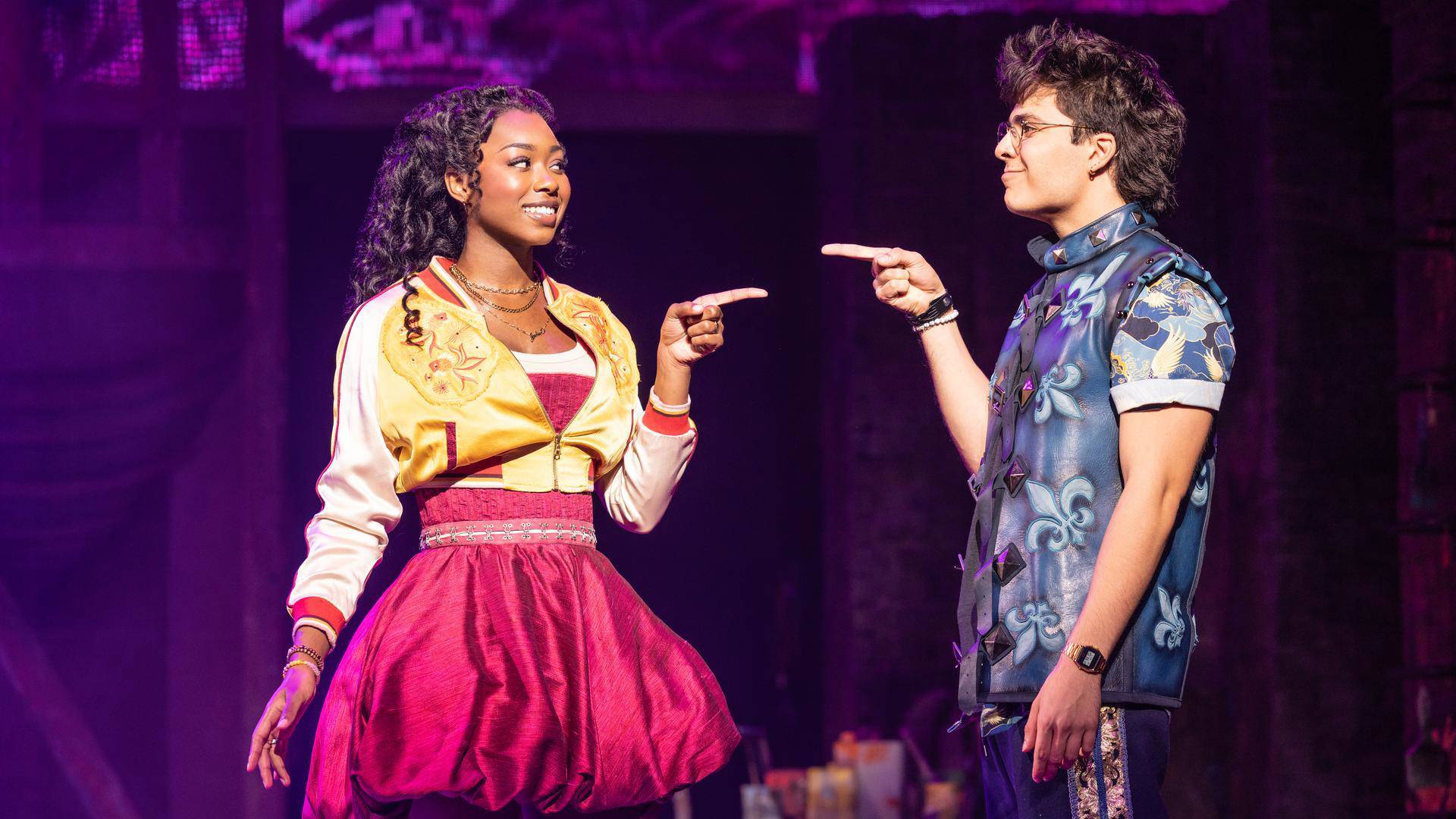 A woman with dark complexion and long dark hair in a yellow bomber jacket and pink dress pointing toward a person with light complexion, medium brown hair and glasses in a blue patterned outfit. They are pointing back at her.