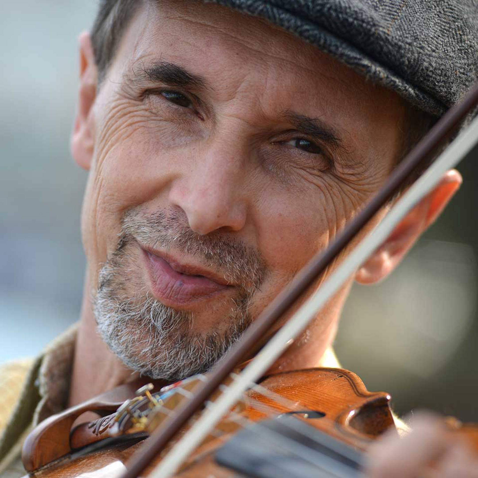 Headshot of a middleaged caucasian man holding a violin. He e has dark eyes and a grey goatee. He is wearing a cap.