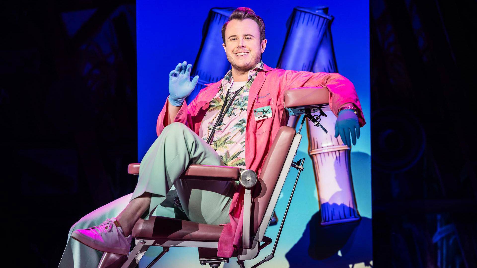 A man with light complexion, brown hair, a pink doctors coat over a Hawaiian shirt, and latex gloves. He is leaning back in an examination chair with his left leg up over the armrest.