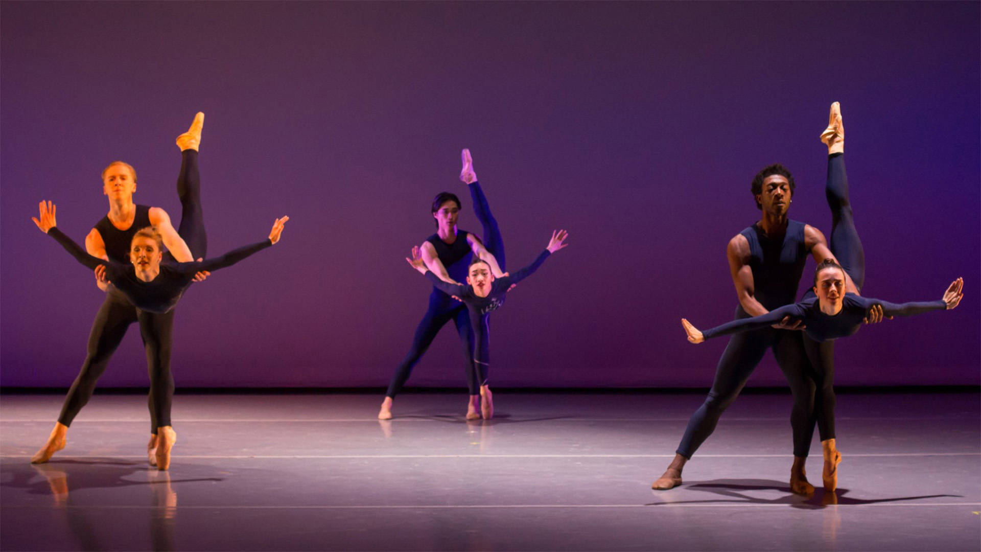 Three pairs of ballet dancers performing on stage with a dark purple background. In each pair, one person is holding the others underarms while they lean forward and lift one back leg up high.