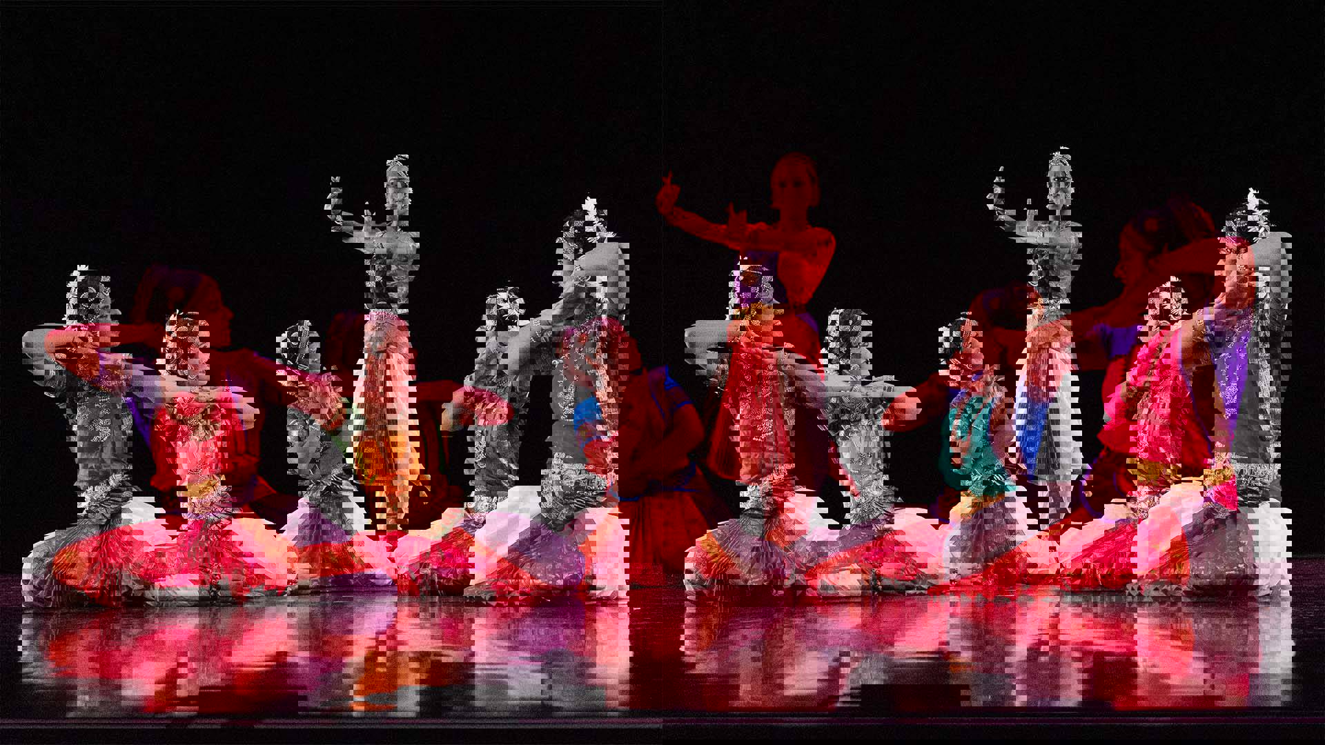 Six women in traditional indian dance outfits performing on a dark stage. Five of them are seated and the sixth stands in the center.