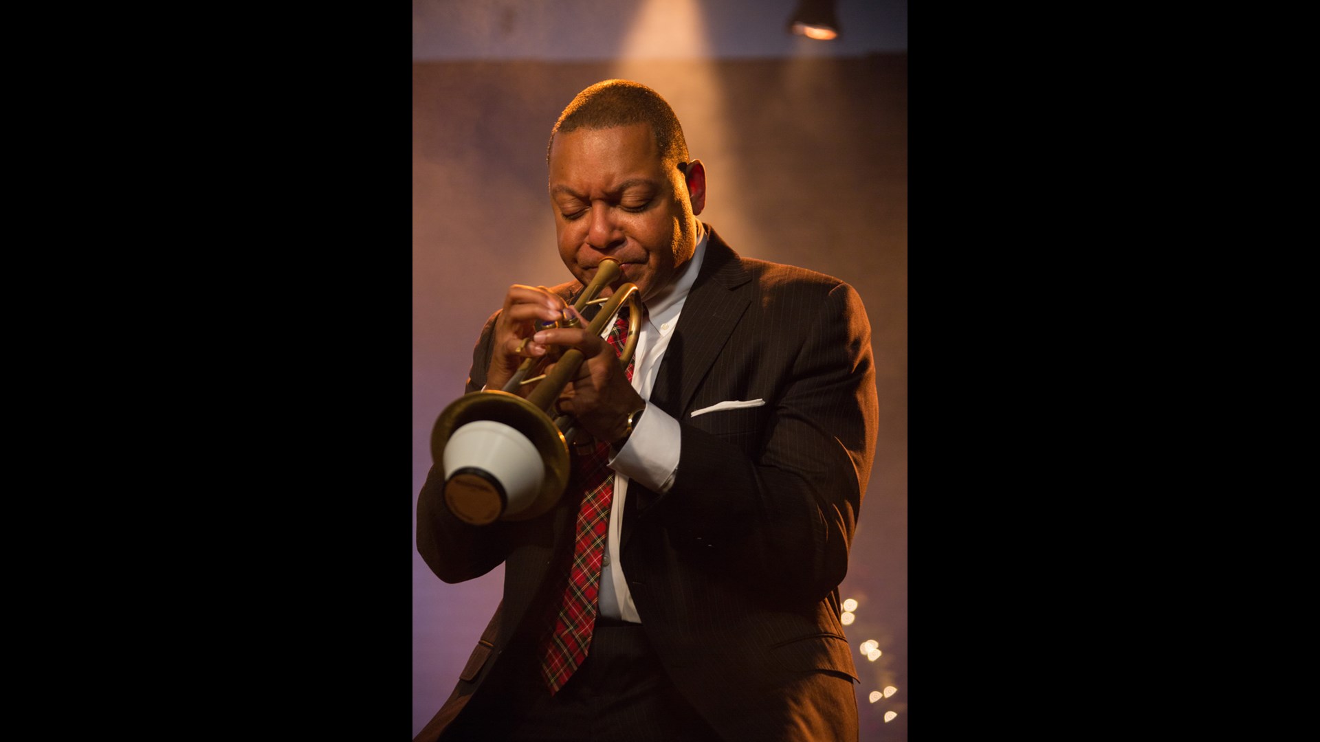 A man with dark complexion and buzzed hair playing trumpet. He is wearing a suit and plaid tie.