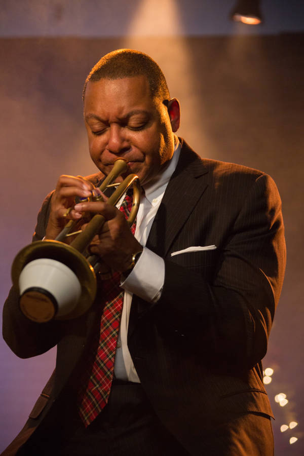 A man with dark complexion and buzzed hair playing trumpet. He is wearing a suit and plaid tie.