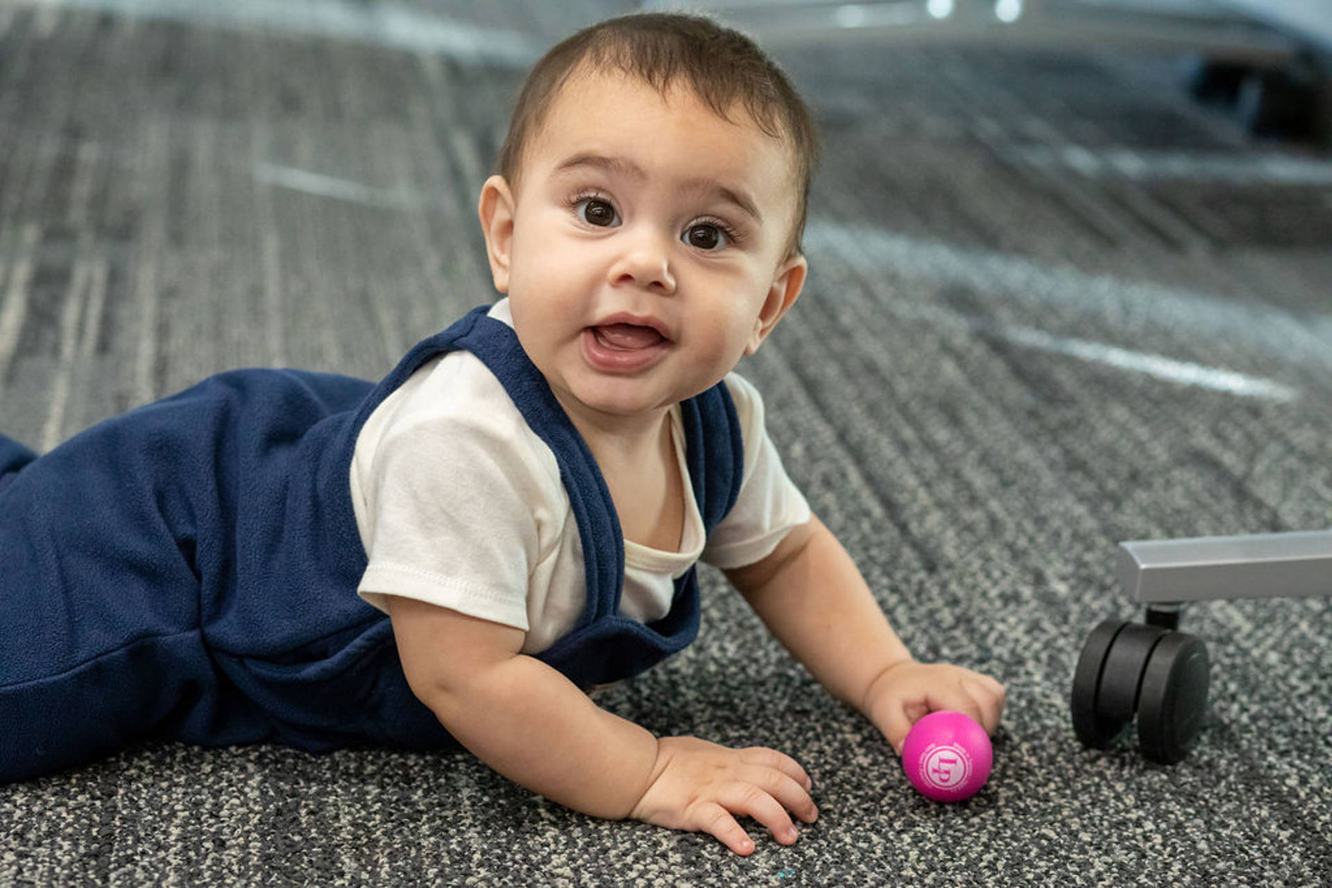 An infant with medium complexion and brown eyes crawling on a carpeted floor and looking straight towards the view. A pink egg shaker is on the ground in front of the infant.