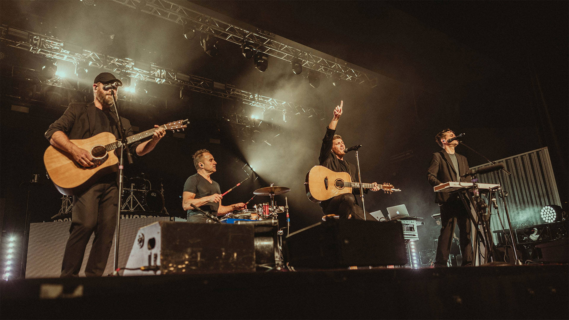 Four men with light complexion playing instruments at the front of the stage and singing into microphones.