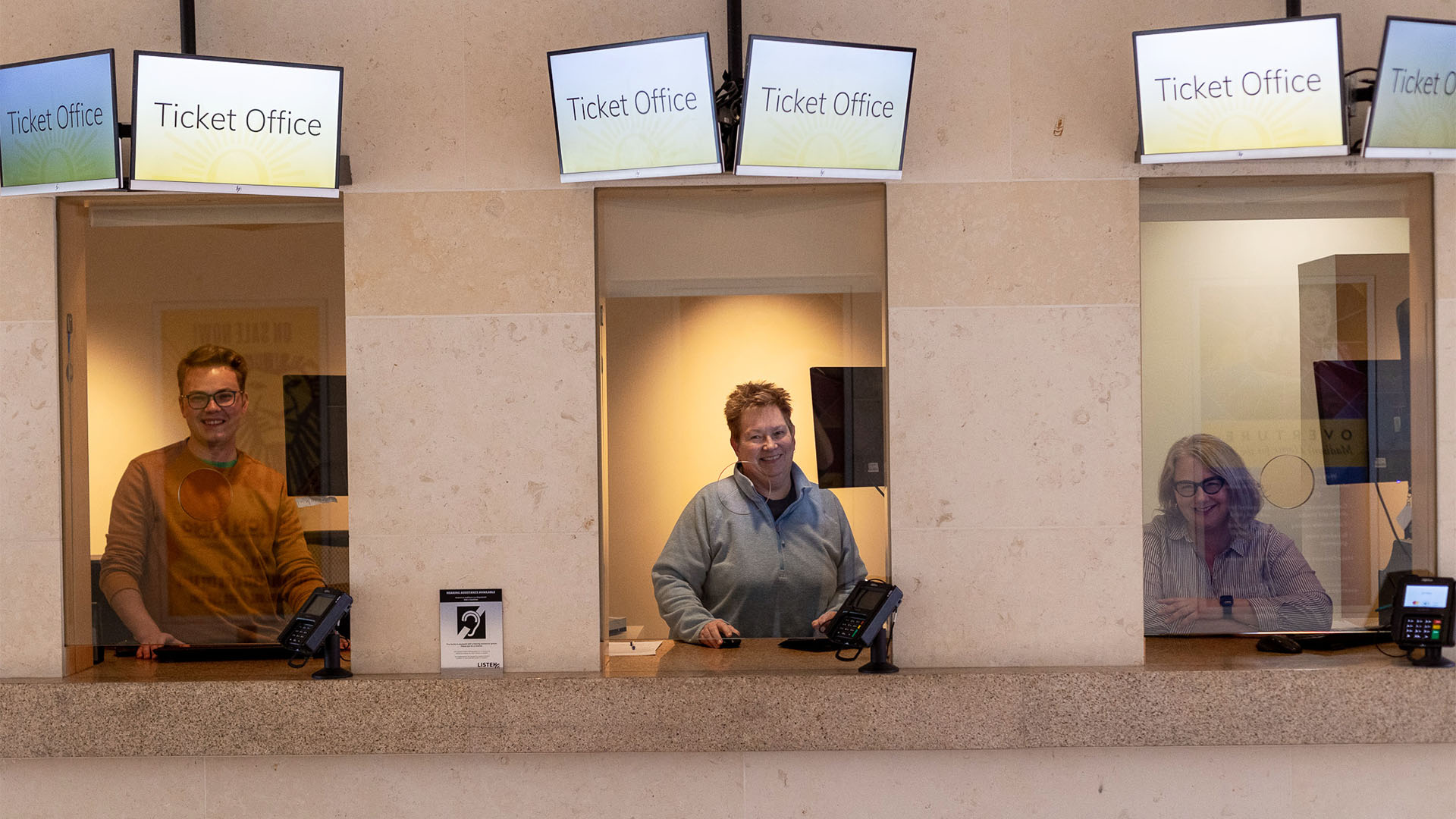 Hugo, a caucasian man in an orange sweater, Mel, a caucasian person in a grey sweater and Melissa, a caucasian woman with medium length grey hair, posing for a photo in Overture's Ticket Office windows.