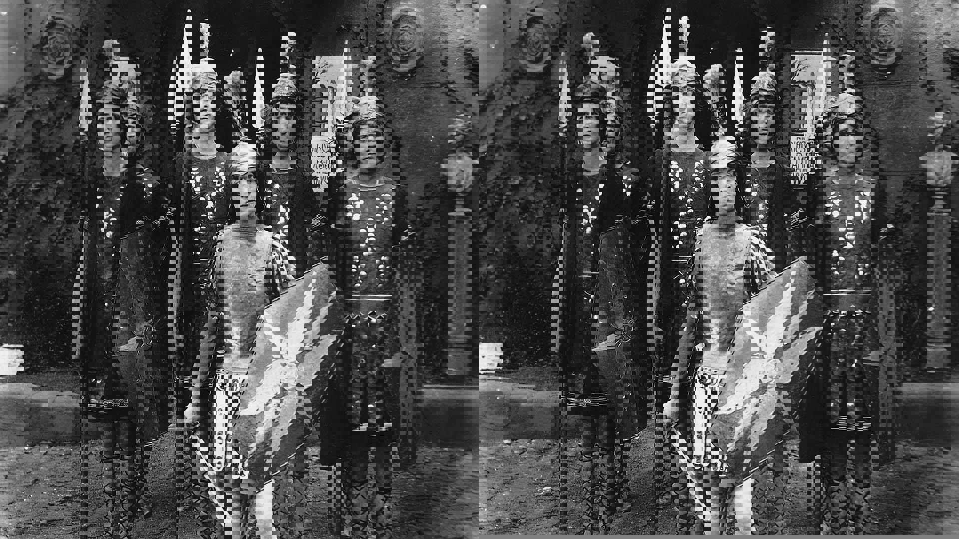 Black and white image of Buster Keaton dress in a Roman warrior outfit with a sword and a shield. He is standing in front of a company of spearmen standing at attention.