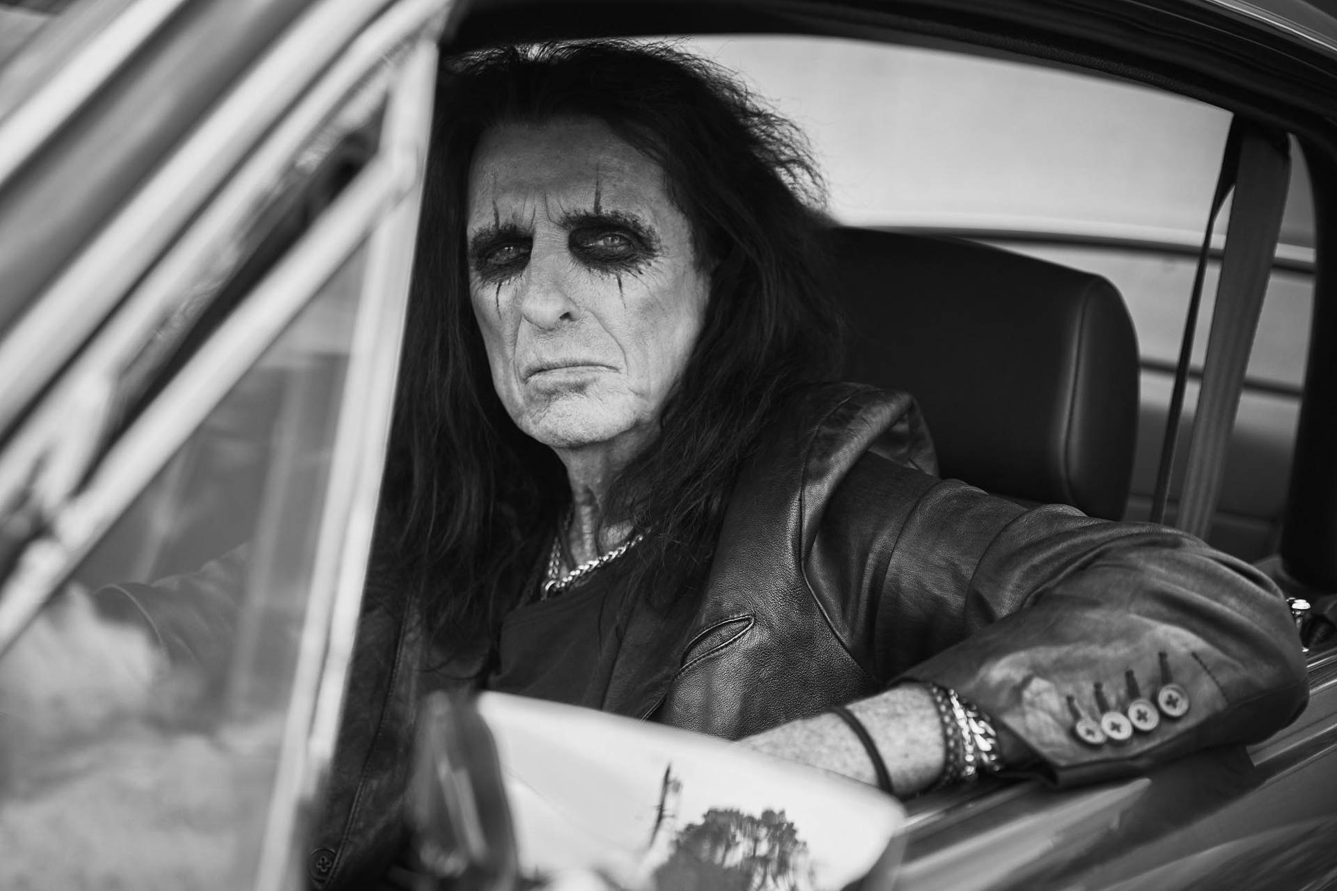 A black and white photo of an older man with long black hair and a lot of eye shadow. He has a black leather jacket and is sitting in a car.