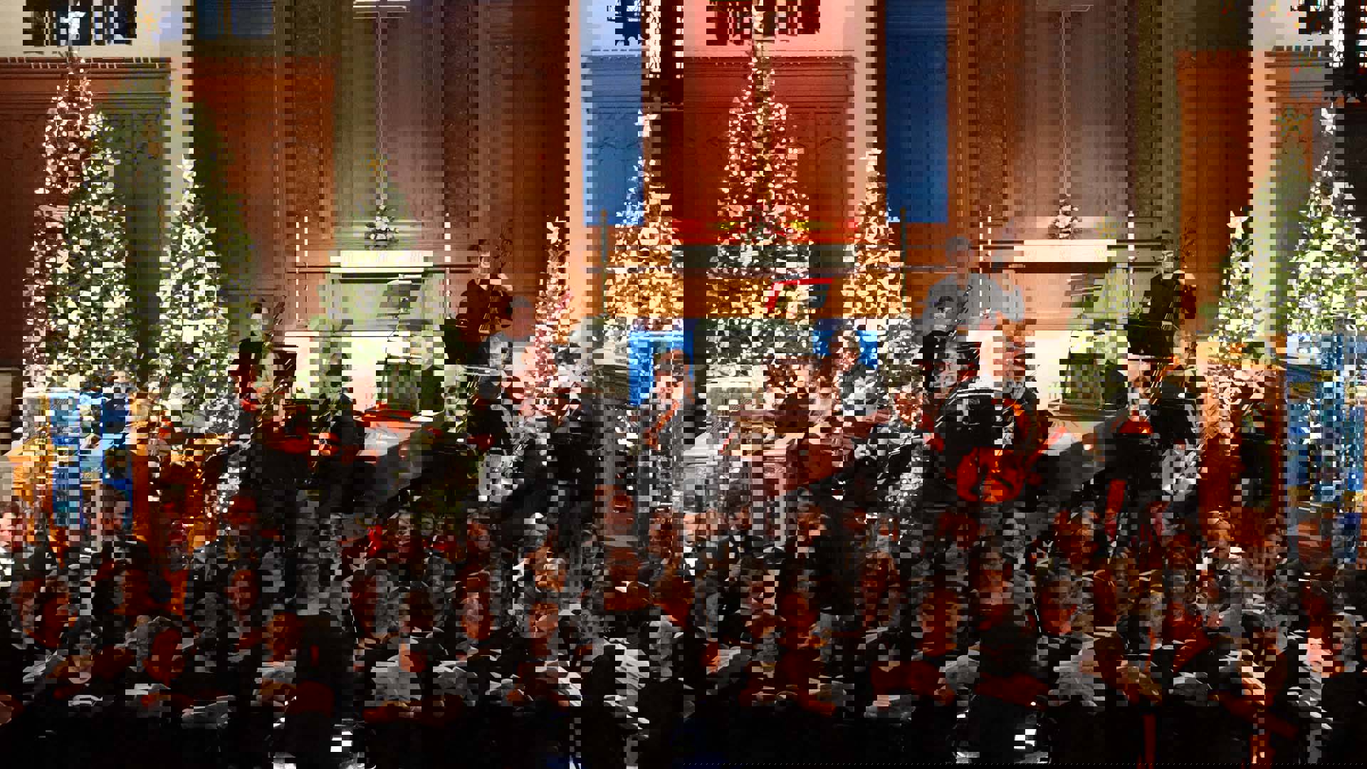 A chamber orchestra and choir dress in black performing in front of a church alter with multiple christmas trees with white ornaments