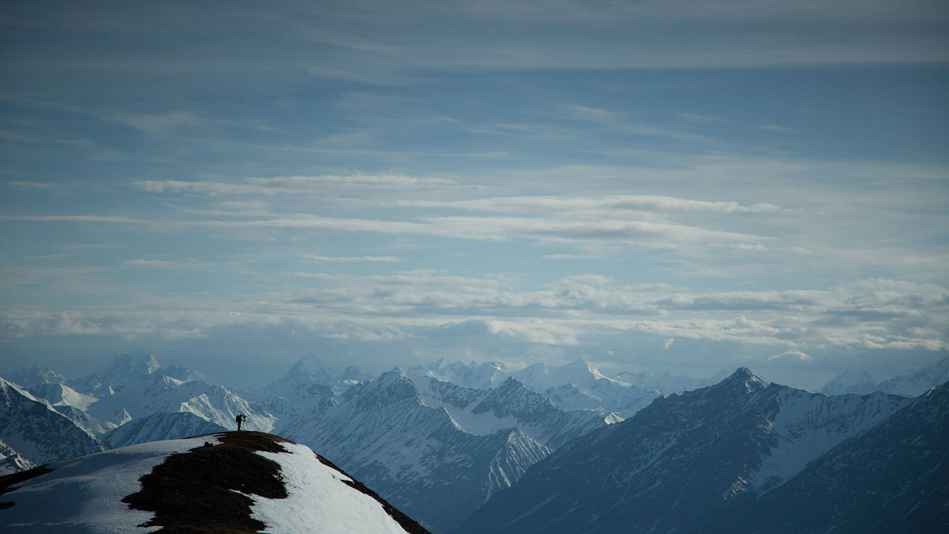 Landscape view of snow-capped mountains in Alaska with cloudy skies. Foreground shows a hill with a photographer's silhouette standing.