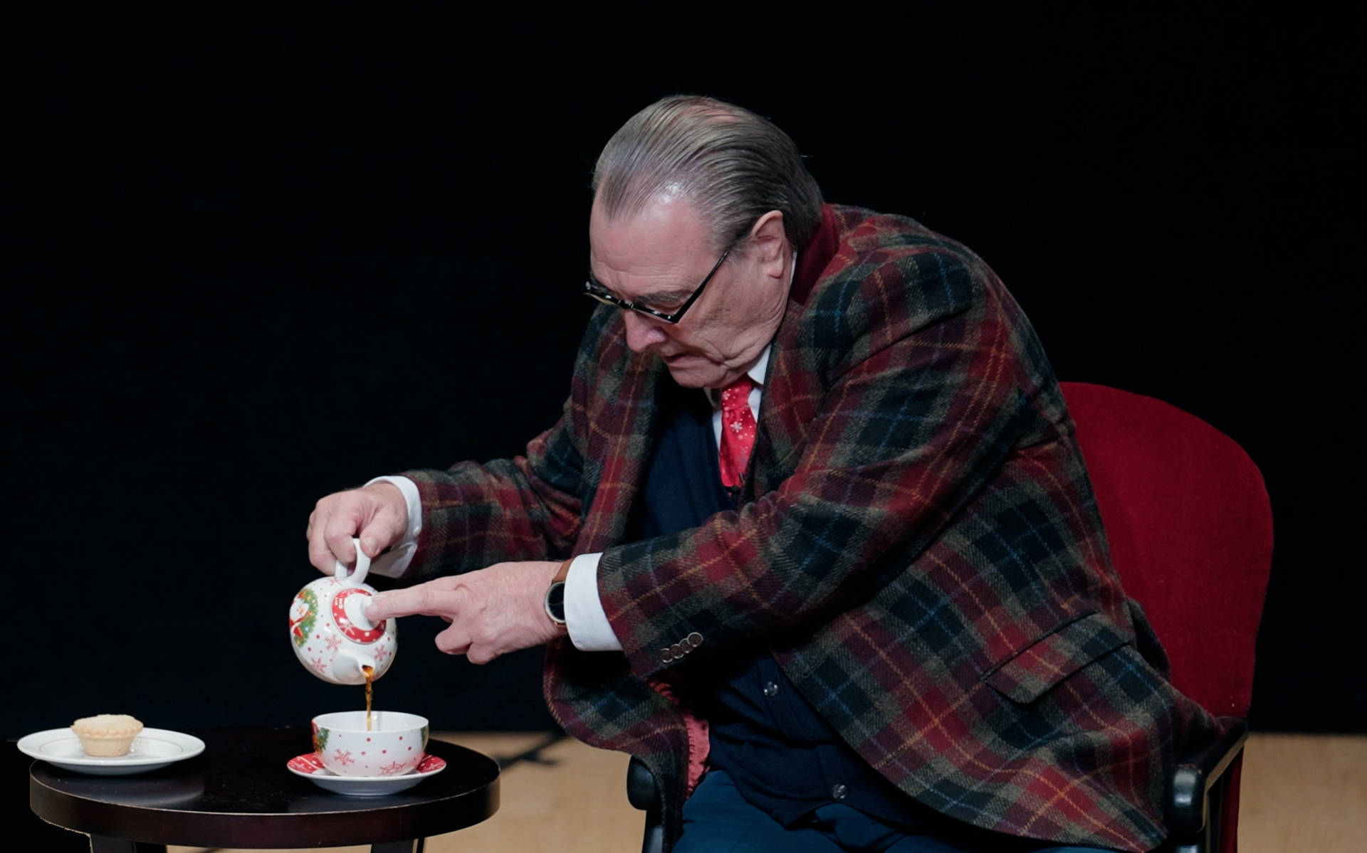 An older, white man with a receding hairline in a blue suit and plaid jacket. He is pouring a cup of tea.