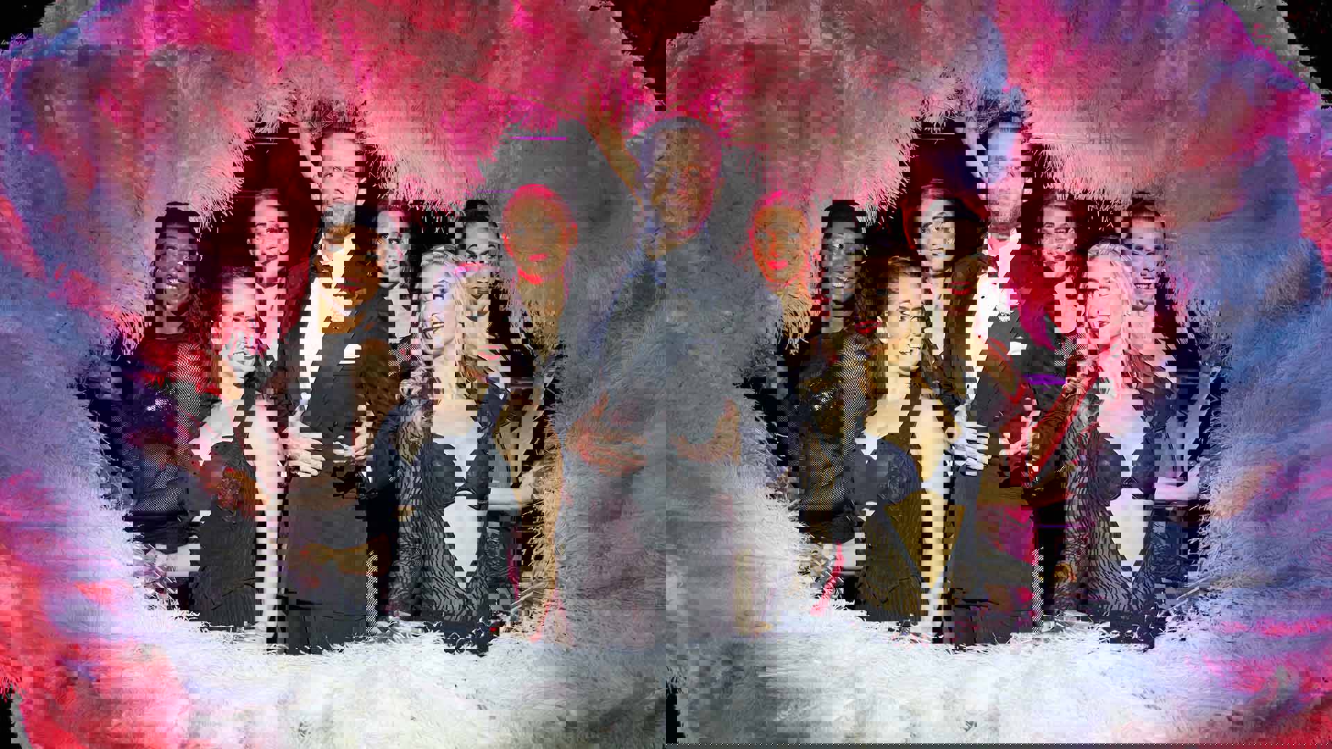 A white man in a tuxedo surrounded by six women in black outfits holding large white feather fans.