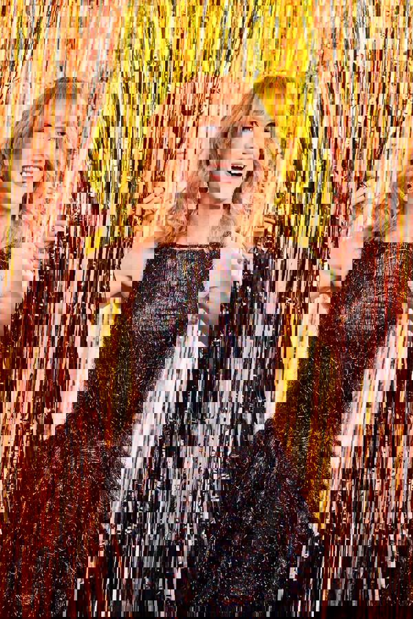 A smiling middle aged woman with light complexion, medium length wavy red hair, and blue eyes. She is wearing a sparkly dark dress and pushing aside hanging reddish gold curtain strips.