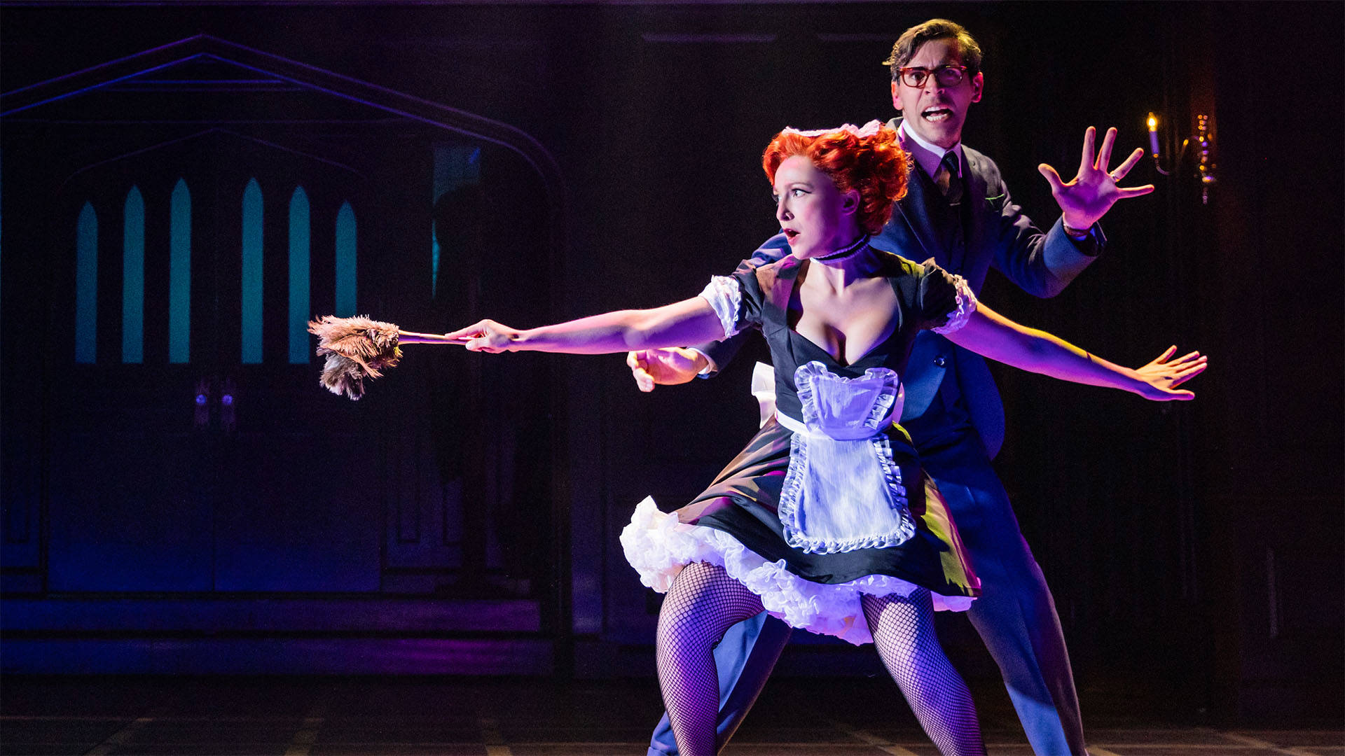 Two actors stand crouched on stage as they are on the lookout for something. A woman in a French maid’s uniform is slowly walking with a feather duster out in front of her as a weapon. A much taller man is behind her looking more frightened than her. 