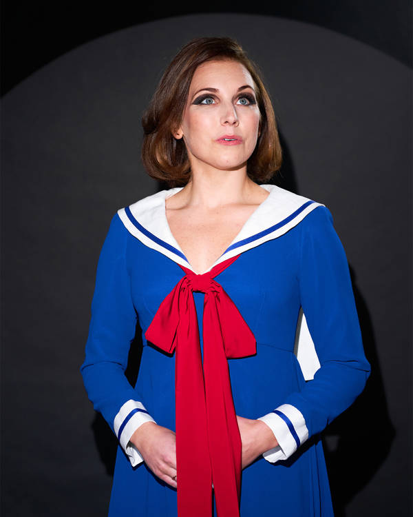 Greyscale headshot of a caucasian woman with medium brown hair and greenish eyes. She is wearing a blue and white top styled like a sailor outfit with a red scarf.