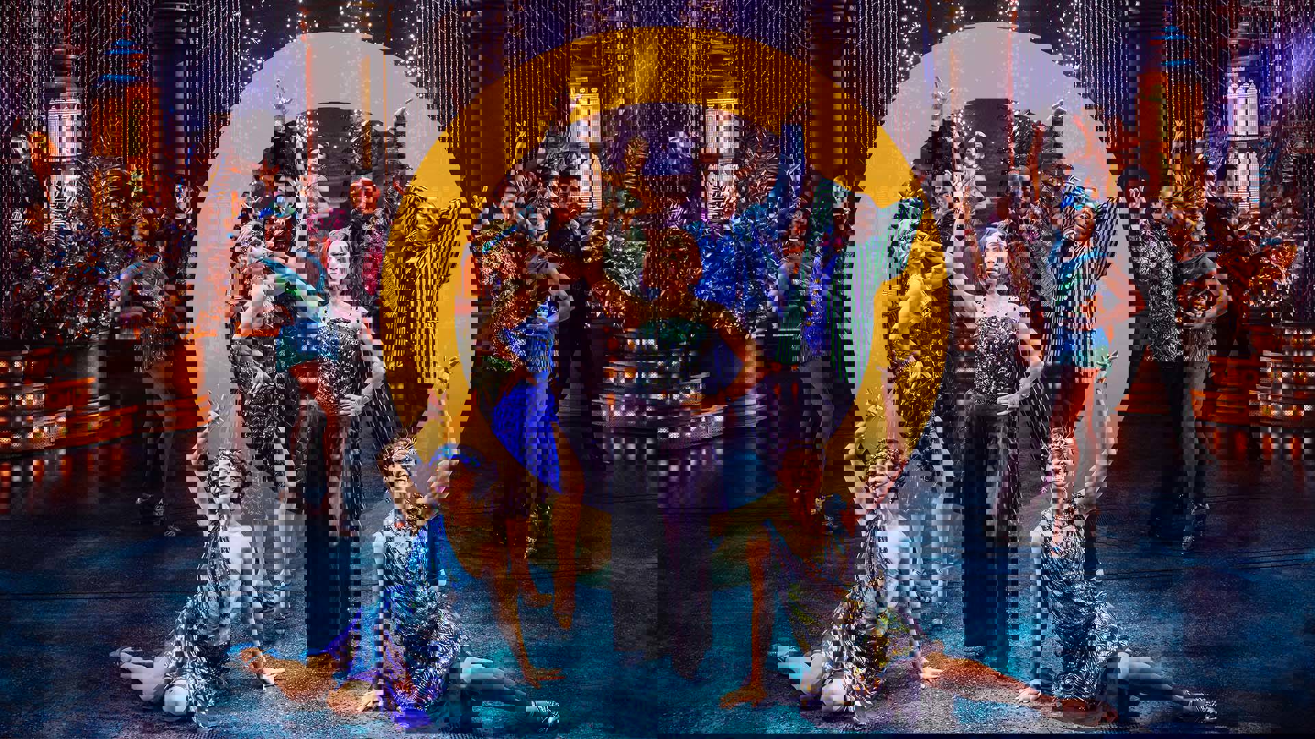 A dazzling stage set-up with art deco decorations as a twelve-person ensemble poses at the center wearing elaborate 1920s party dresses and suits, all in teals, blues and purples. A large yellow "O" has been edited into the center of the image.