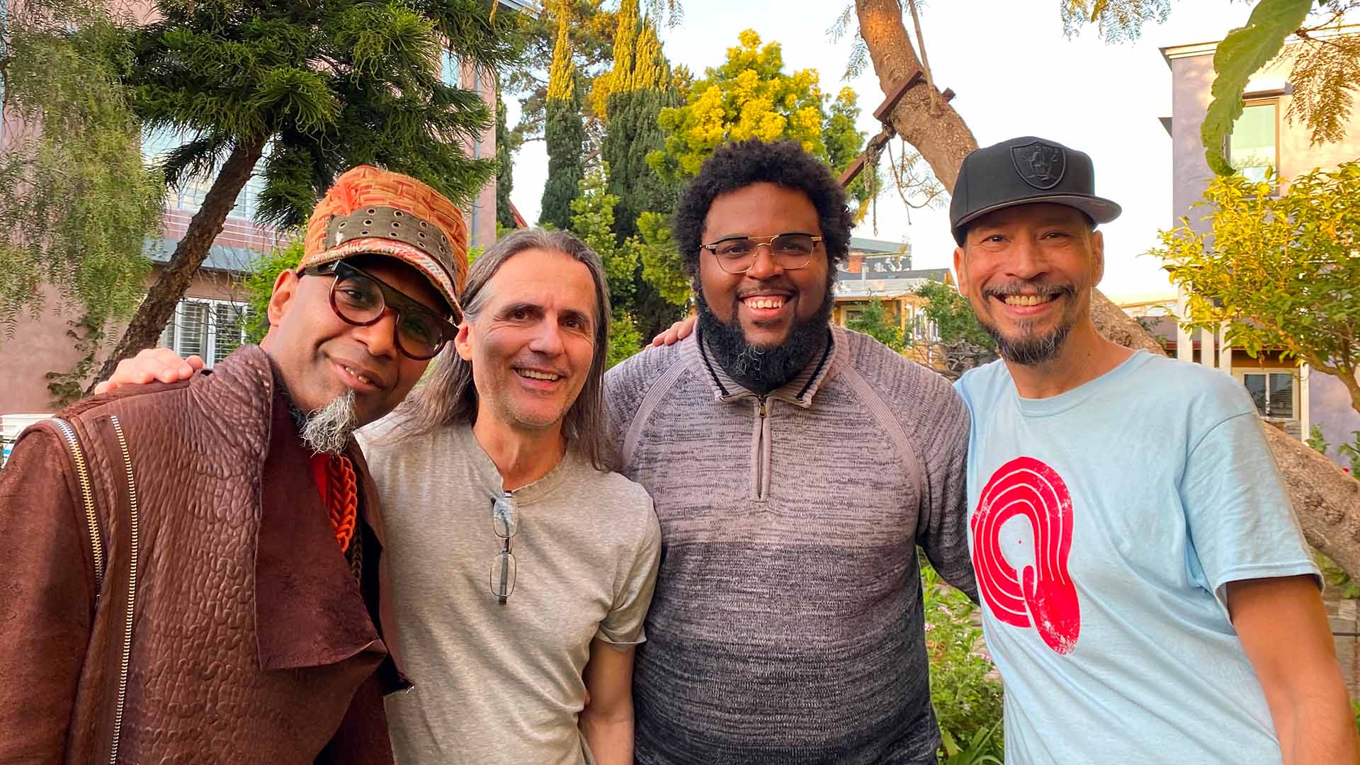 Four men with kind smiles and colorful clothing stand together for a picture. They are outdoors.