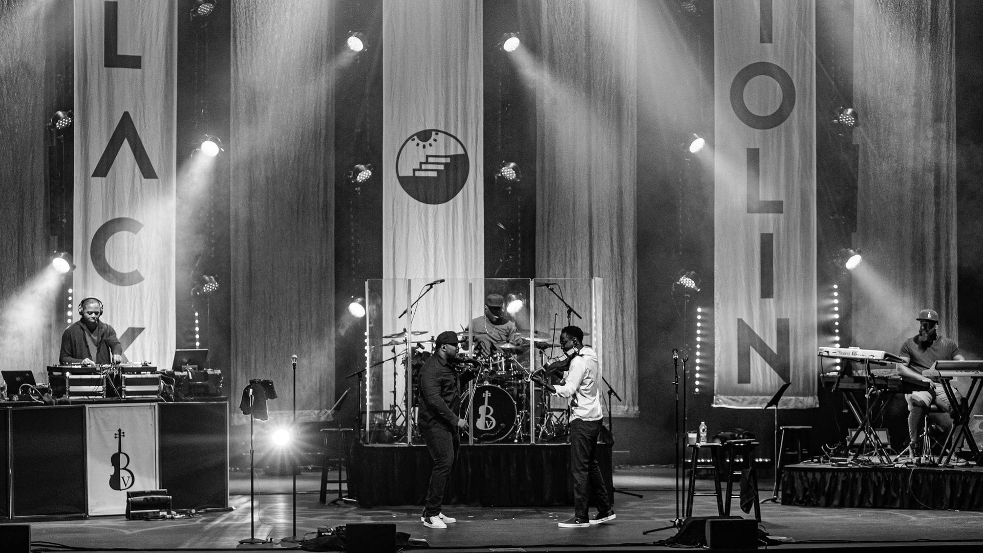Grayscale shot of Black Violin in performance. Two violin players are at the front. A keyboard player, DJ and drummer are behind.