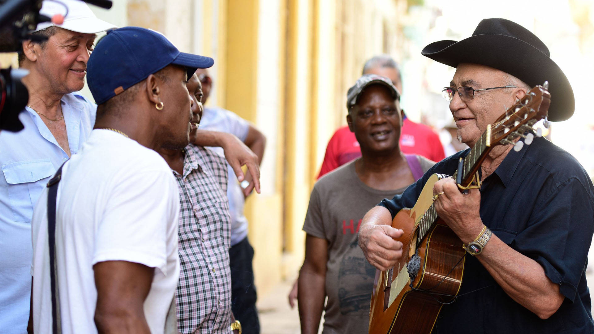 Eliades Ochoa strumming a guitar in front of a small crowd of dark skin toned people in a street.
