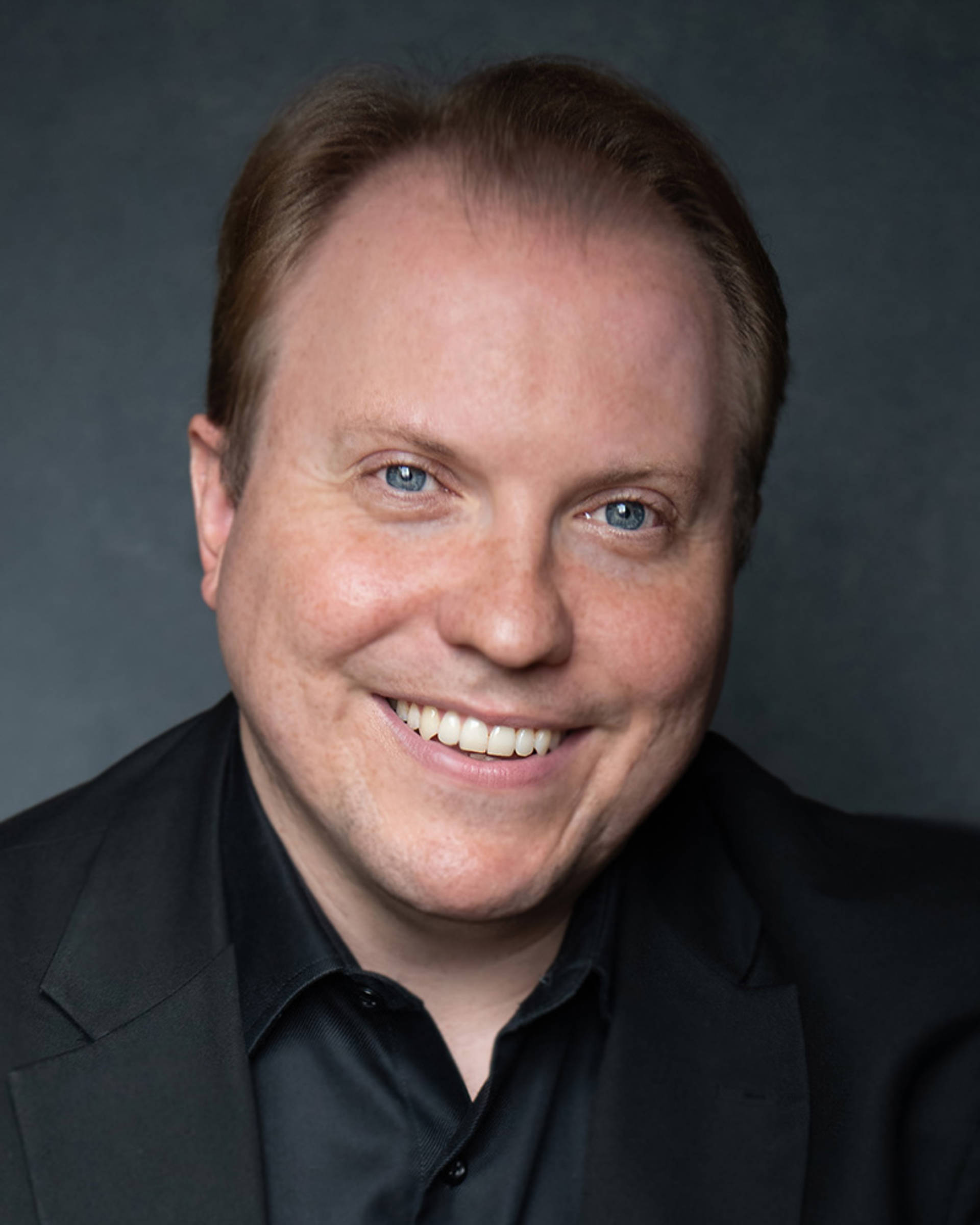 Headshot of a caucasian man with short dark blonde hair and blue eyes. He is wearing a black collared shirt and jacket.