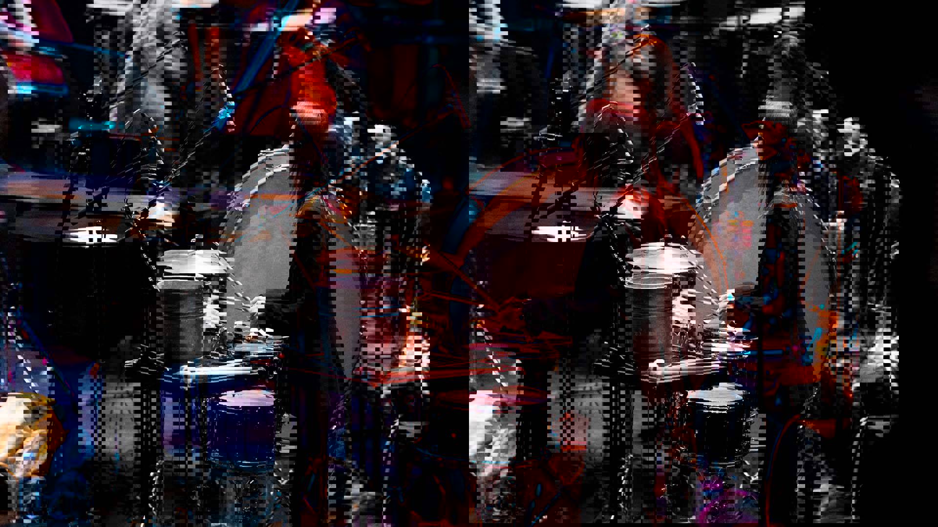 A percussionist with long brown hair and a black suit hitting a hi-hat while performing on stage.