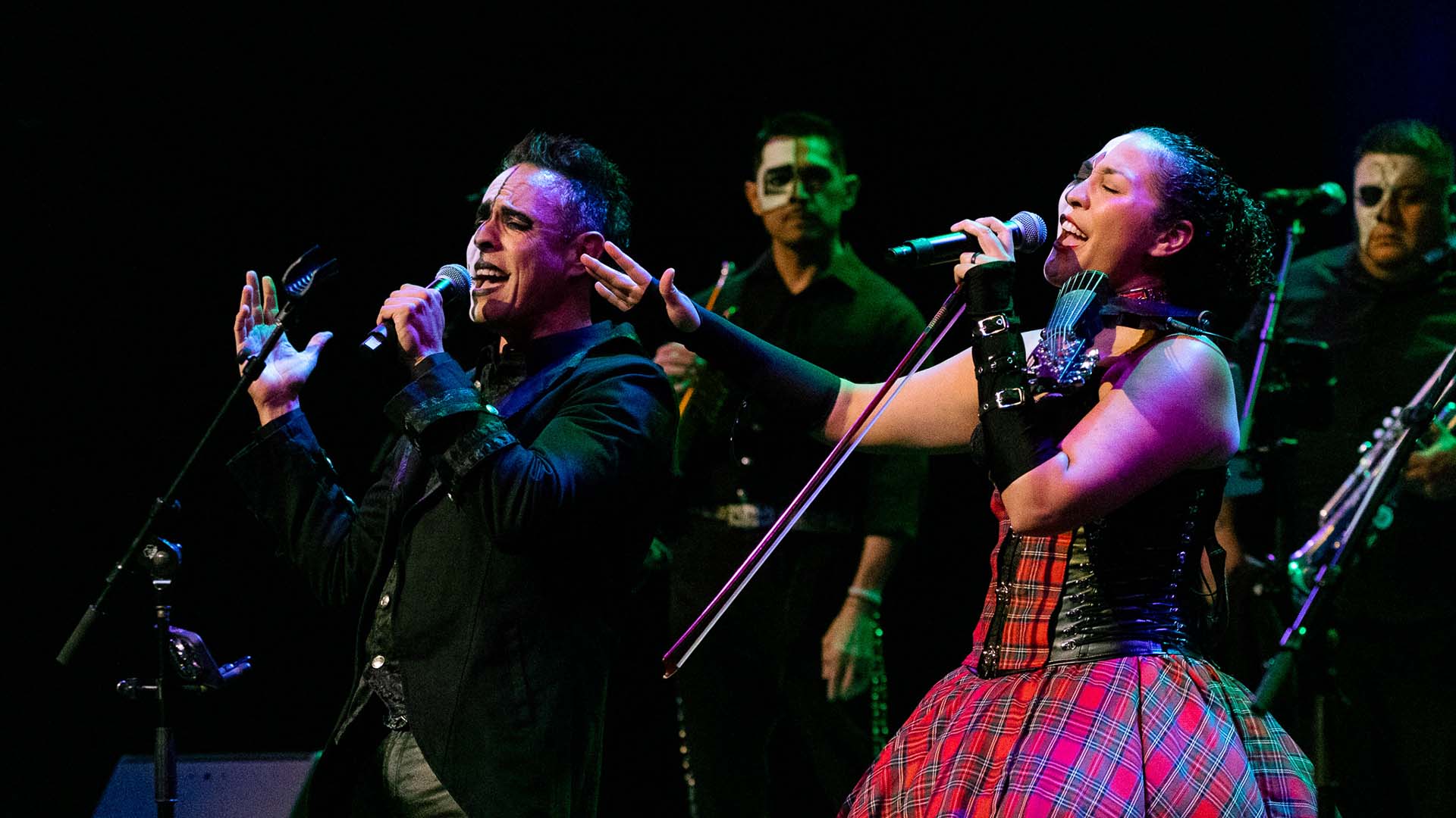 A man and woman with medium complexion and black and white face paint sing passionately into microphones and gesture toward the stage.
