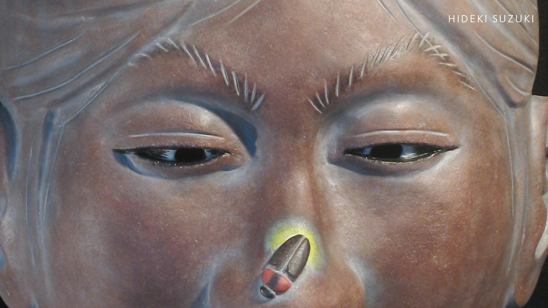 A close up of a dark brown clay mask of a person's face. Their eyes are looking down at their nose where a beetle sits with bright green coloring underneath.